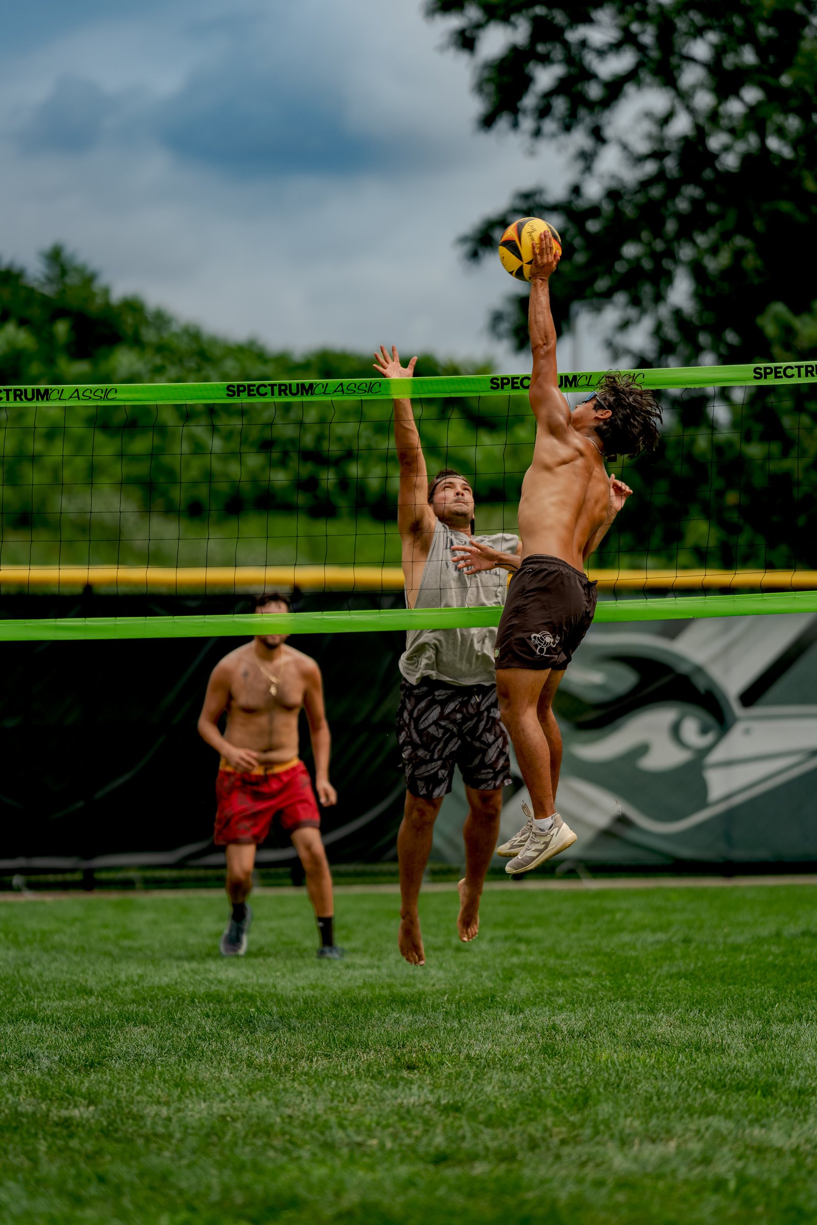 A player in brown shorts spikes a volleyball over the net while another player in a gray shirt attempts to block, with a player in red shorts in the background.