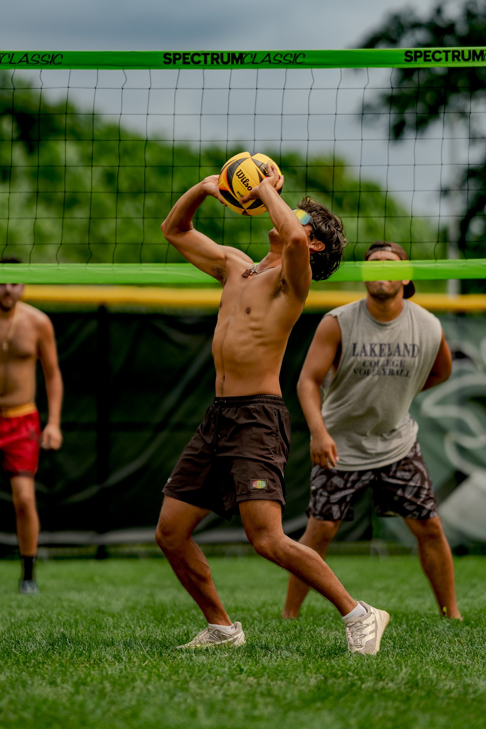 A player in brown shorts prepares to serve a volleyball over the net, with two other players visible in the background on a grassy court.