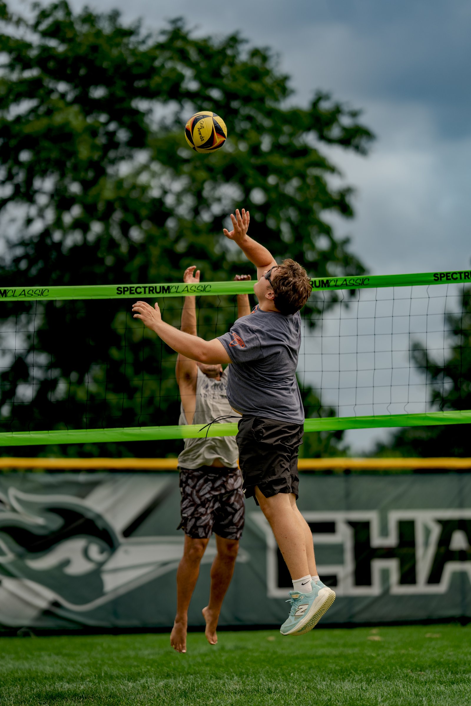 A player in a gray shirt jumps to hit a volleyball over the net, with another player in a gray tank top behind him.