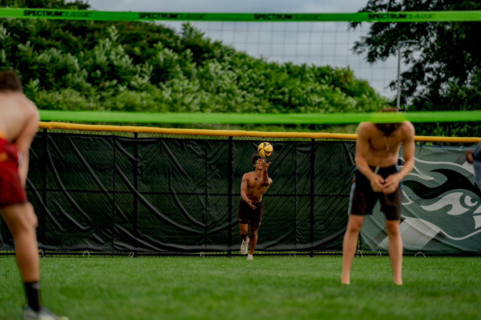 A player in brown shorts and a player in red shorts are on the court, with a player in black shorts tossing a volleyball.