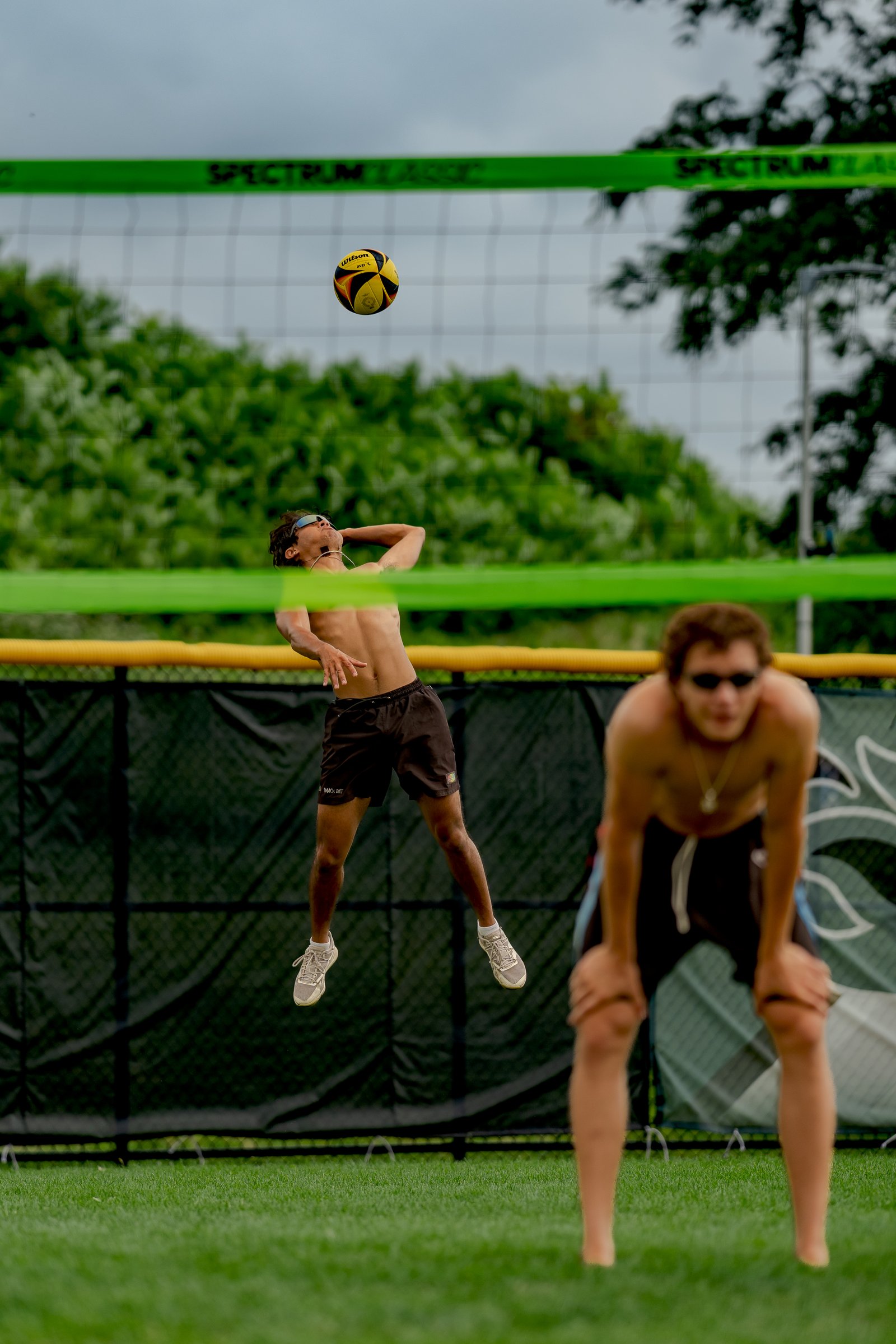 A player in black shorts jumps to hit a volleyball over the net, with another player in black shorts in the foreground.