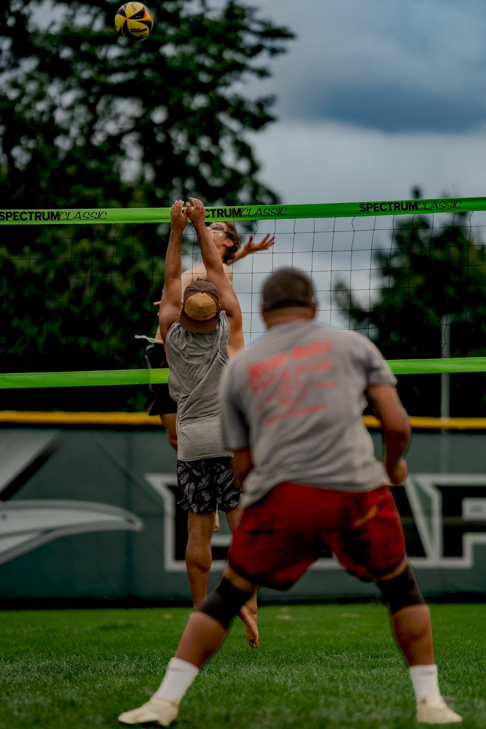 A player in a grey top and patterned shorts reaches for the ball over the net, with another player in red shorts in the foreground.