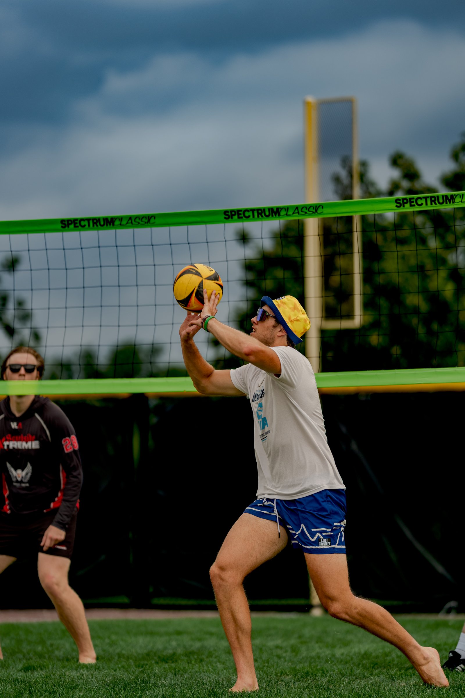 A player in a white shirt and blue shorts sets a volleyball over the net, with another player in a black and red jersey visible.