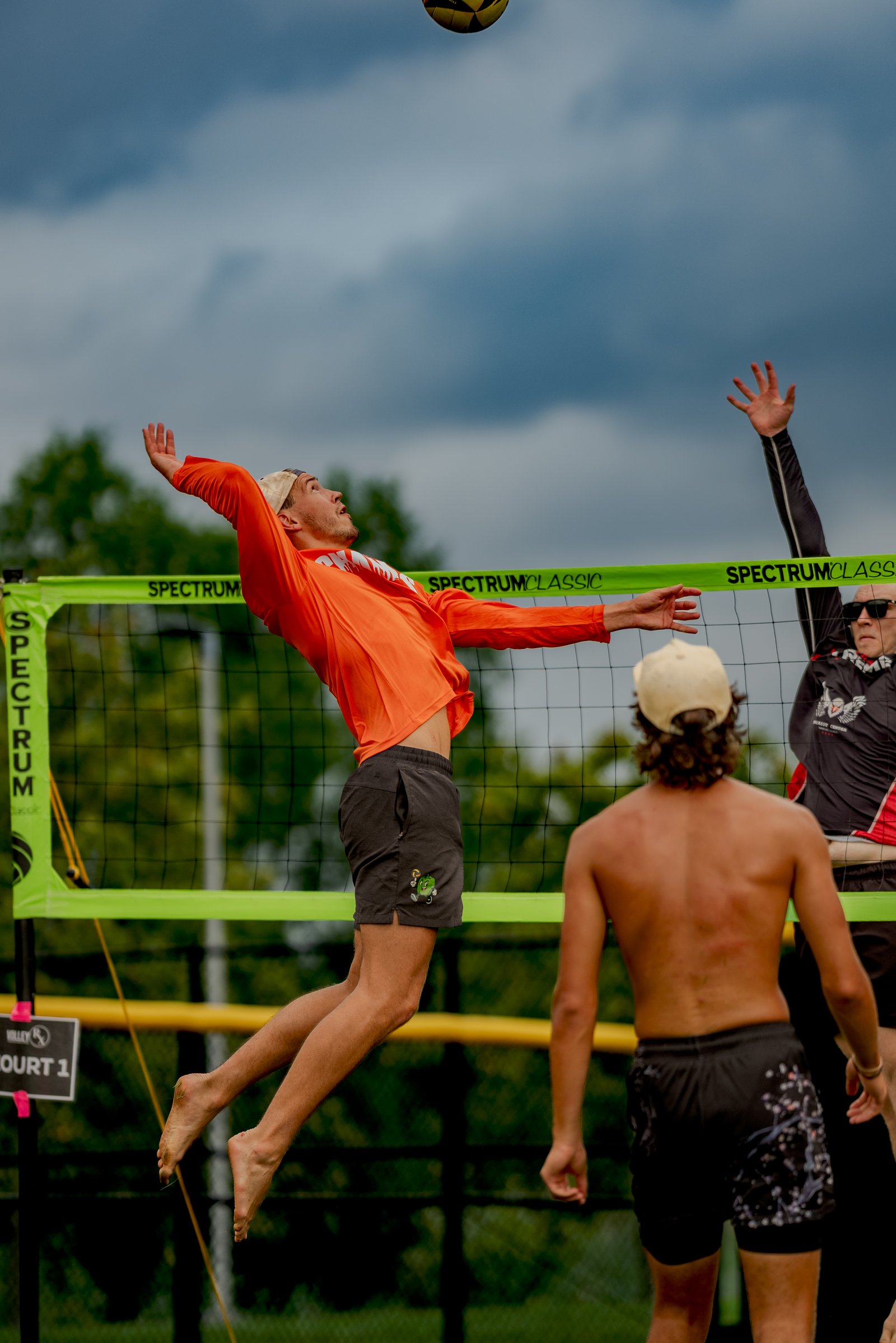 A player in orange jumps to hit a volleyball over the net, with another player in black and red reaching up to block.