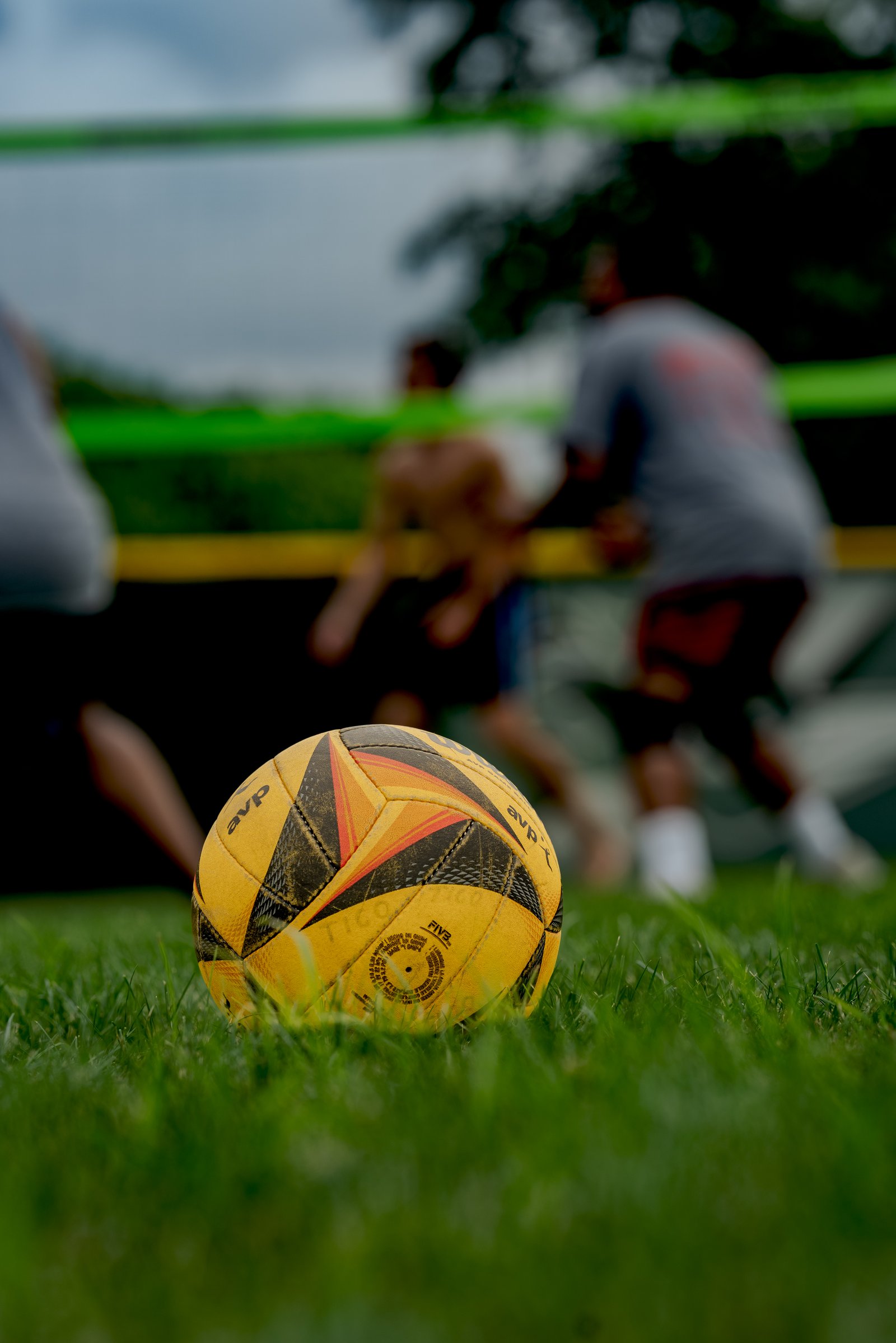 A volleyball rests on grass as players in gray and red shorts move on the court behind a green net.