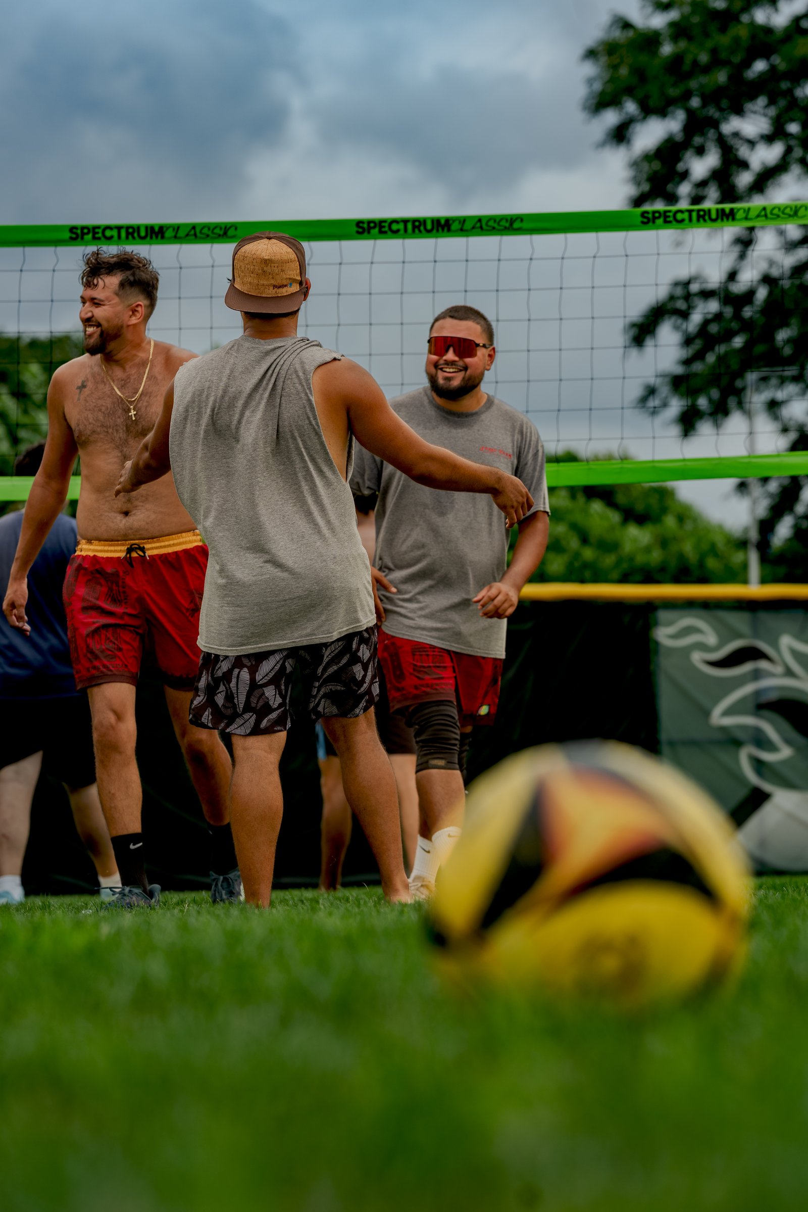 Players in red shorts and grey tops interact near a volleyball net, with a ball in the foreground.