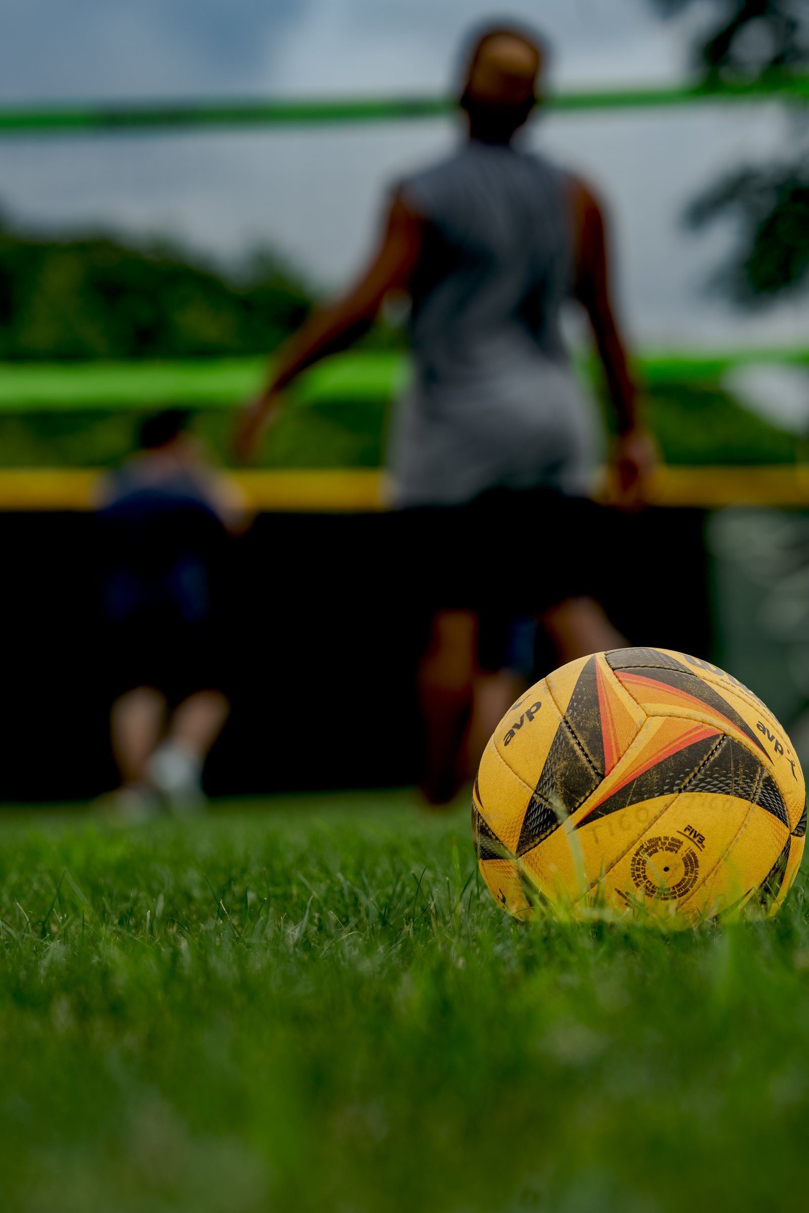 A yellow volleyball rests on grass with blurred players in the background near a net.