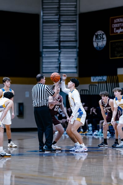 A referee holds a basketball as a player in white and blue jumps. A player in maroon stands nearby.
