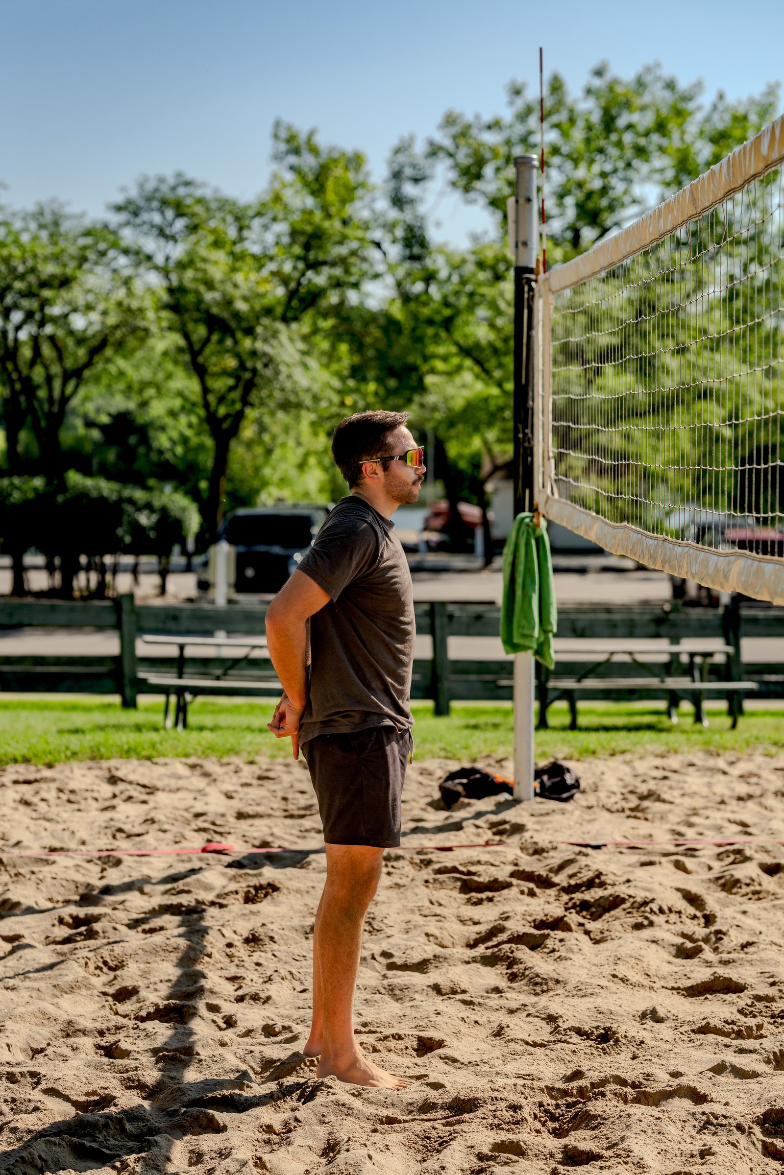 A player in a dark grey shirt and dark shorts stands on a sand court near a volleyball net, stretching.