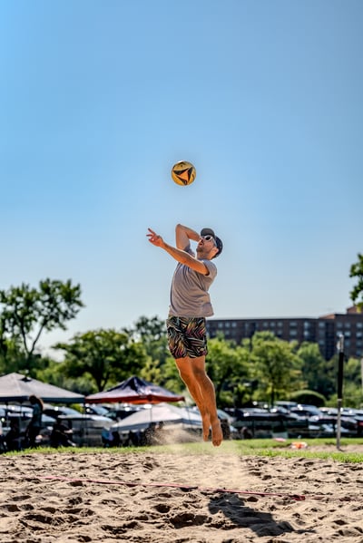 A player in a grey shirt and patterned shorts jumps to hit a volleyball on a sunny day at the beach.