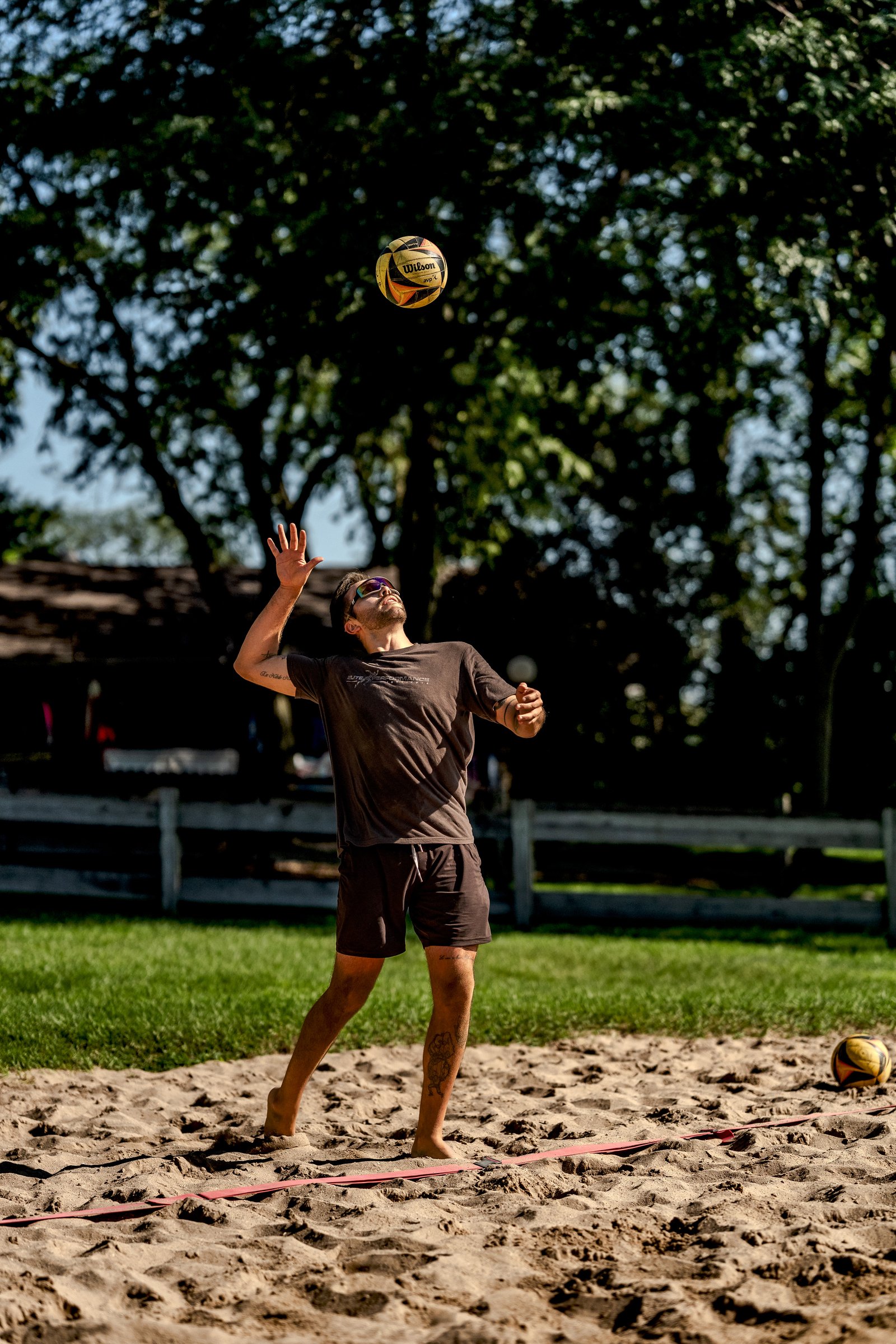 A player in a dark grey shirt and dark shorts serves a volleyball on a sand court with trees in the background.