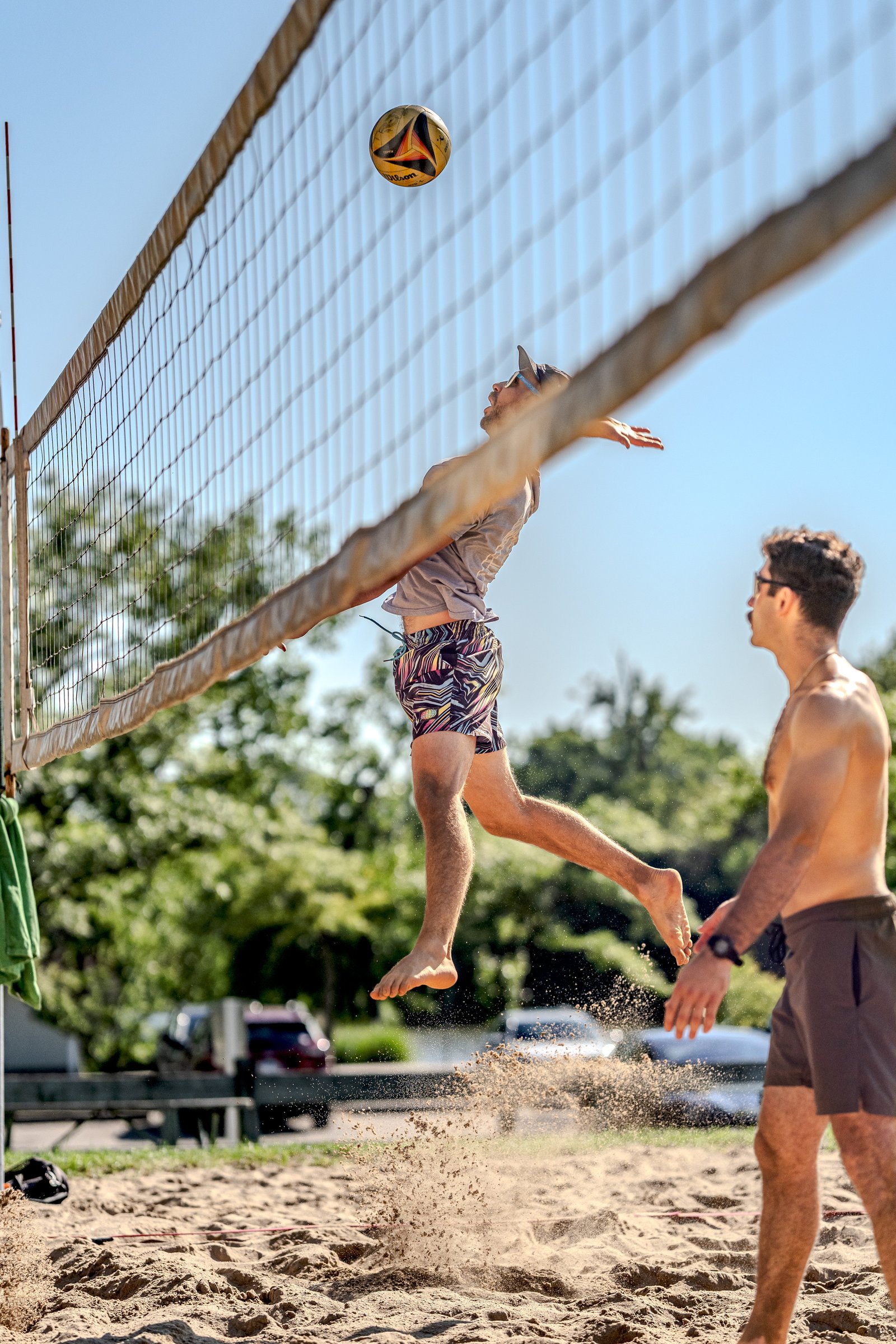 A player in patterned shorts jumps to hit a volleyball over the net, with another player in brown shorts watching.