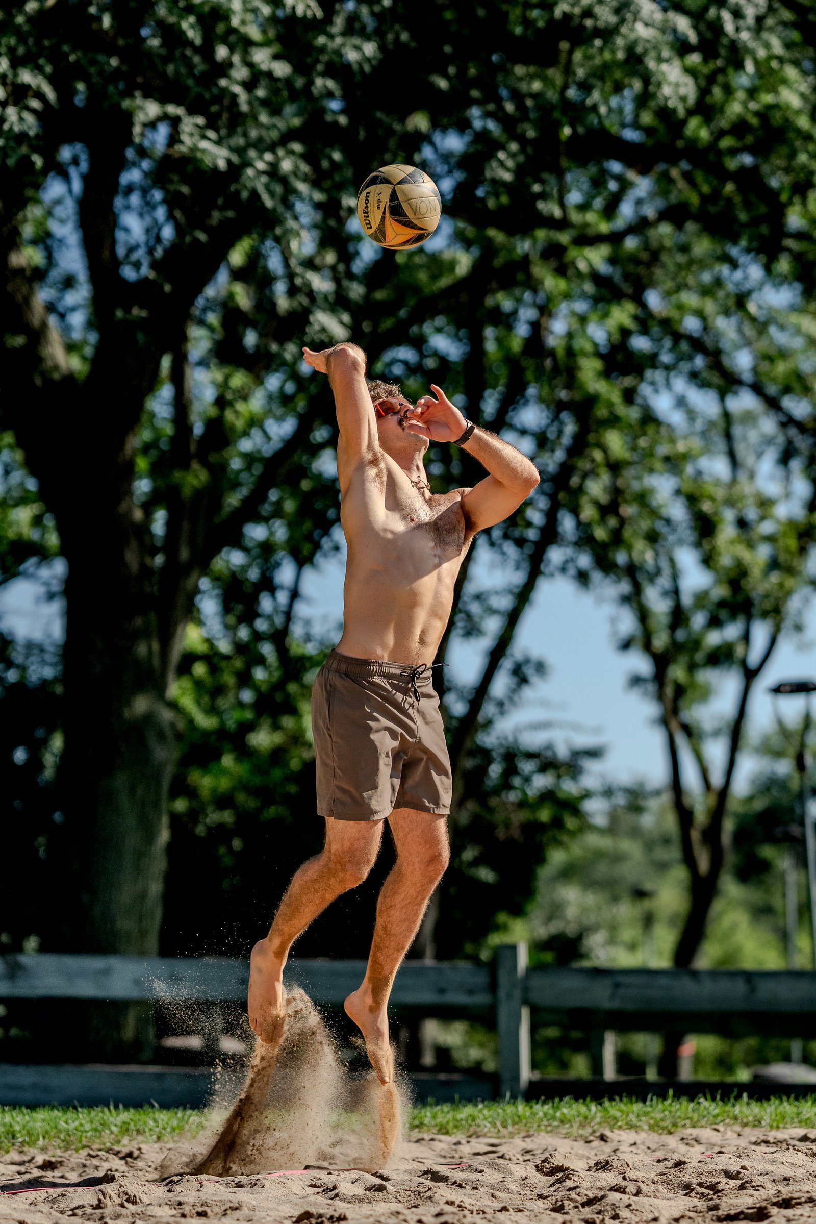 A player in brown shorts jumps to hit a volleyball in the air on a sandy court.