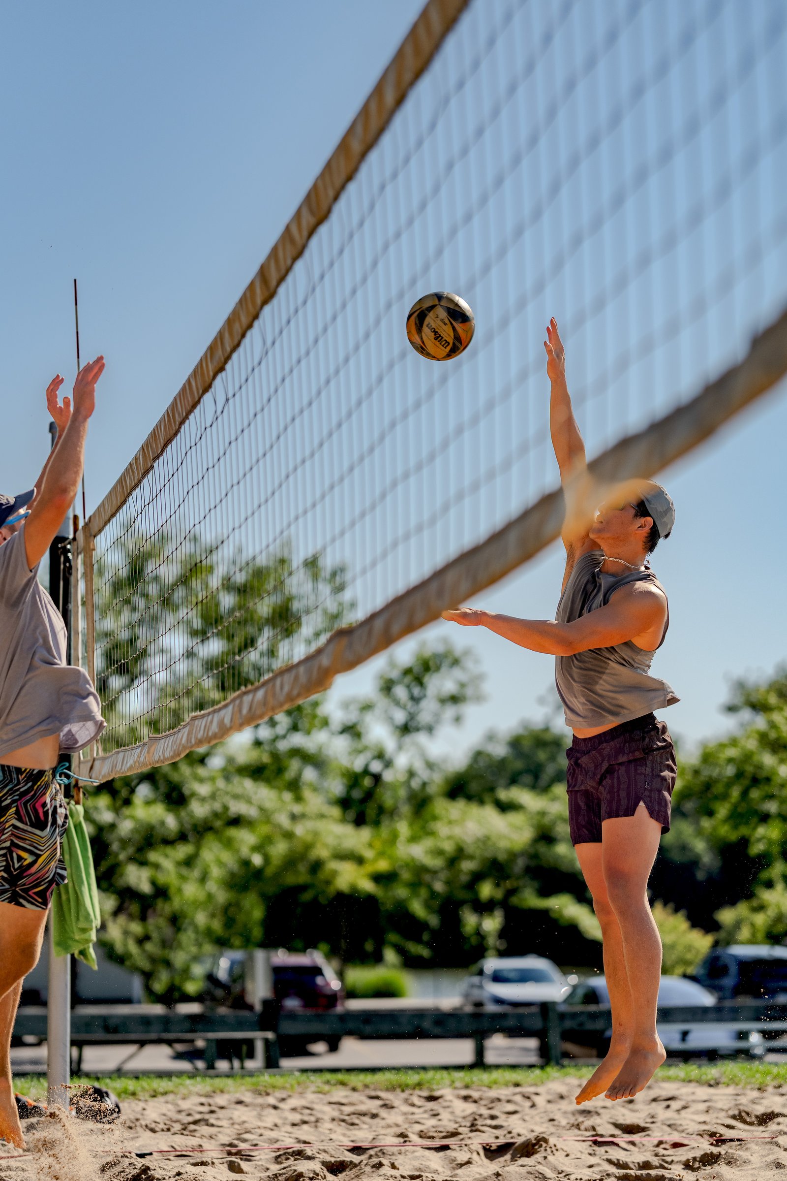 A player in a gray top and dark shorts jumps to hit a volleyball over the net, with another player in a gray top and patterned shorts nearby.
