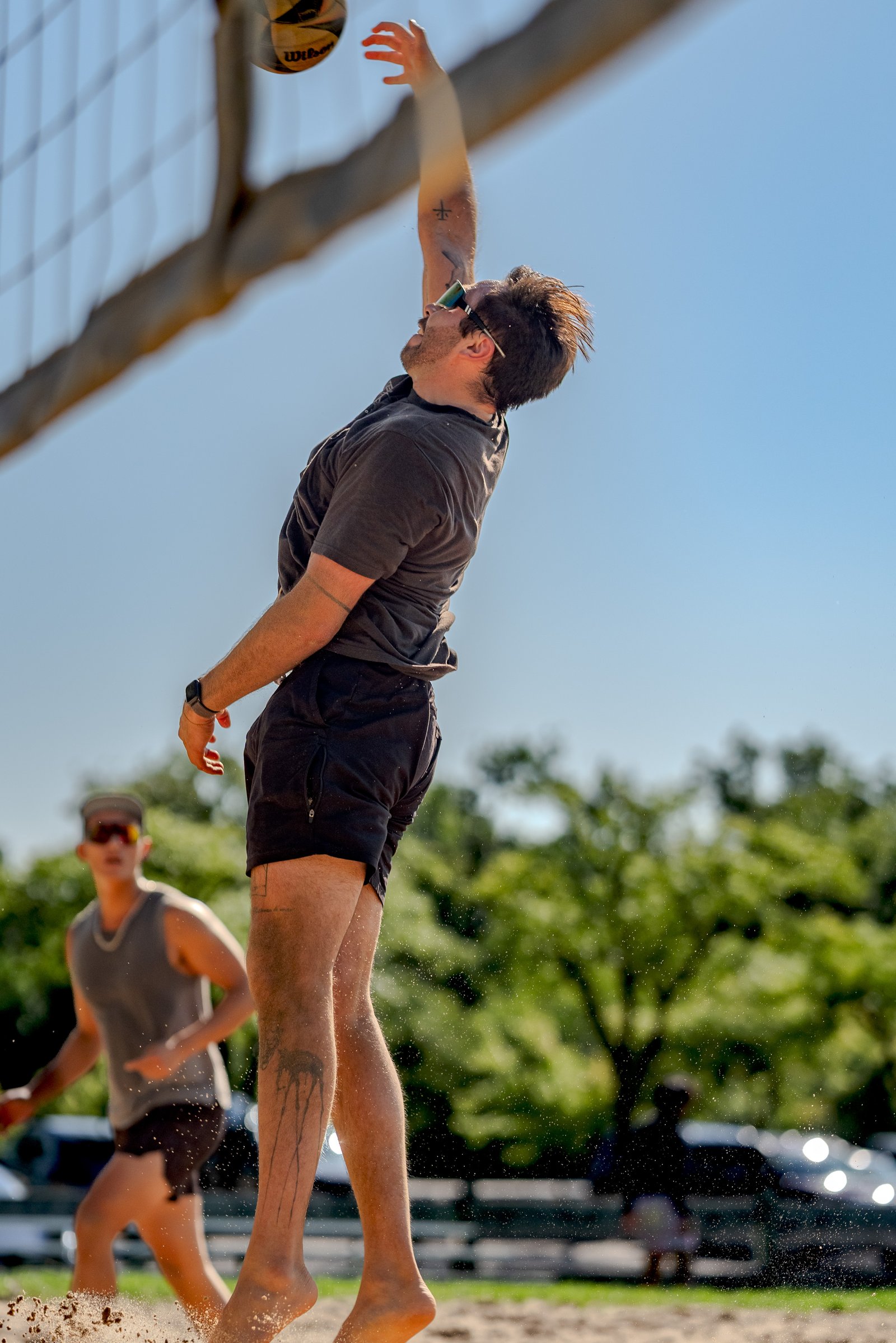A player in a dark grey shirt and black shorts jumps to hit a volleyball over the net, with another player in a grey tank top in the background.