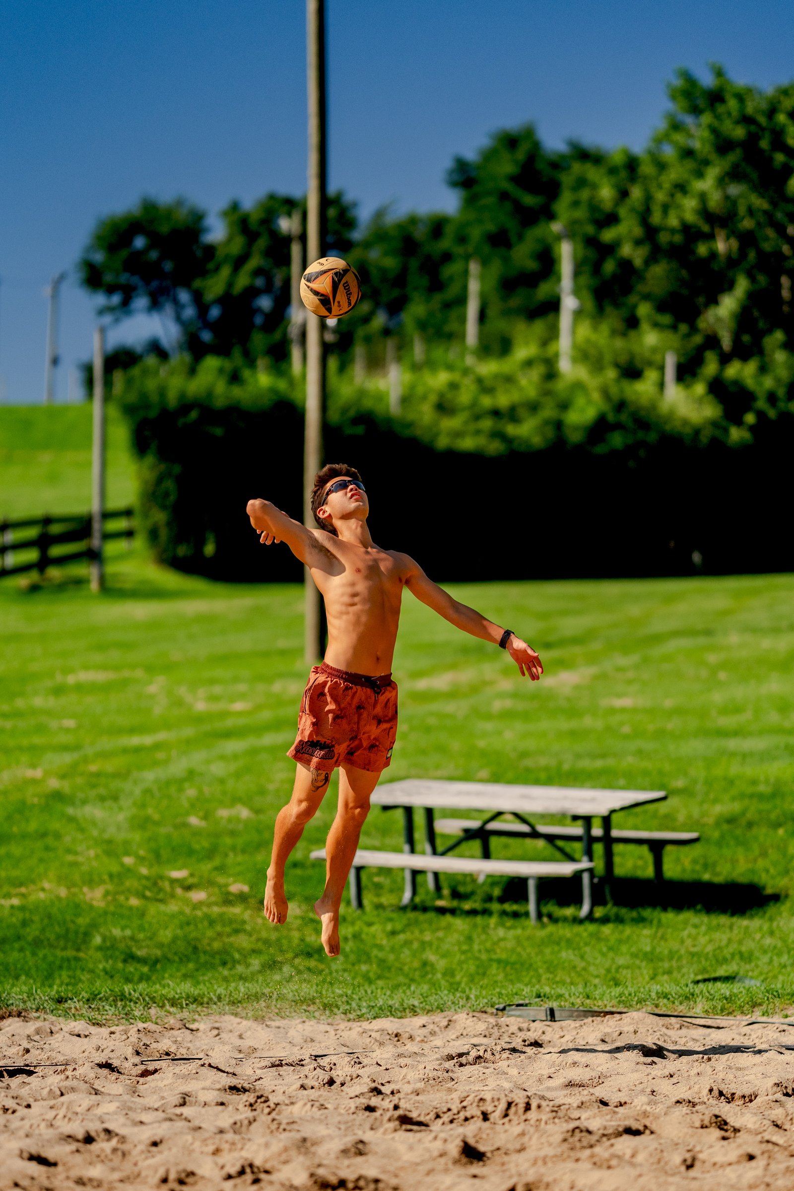 A player in orange shorts jumps to hit a volleyball on a sand court with a picnic table and trees in the background.