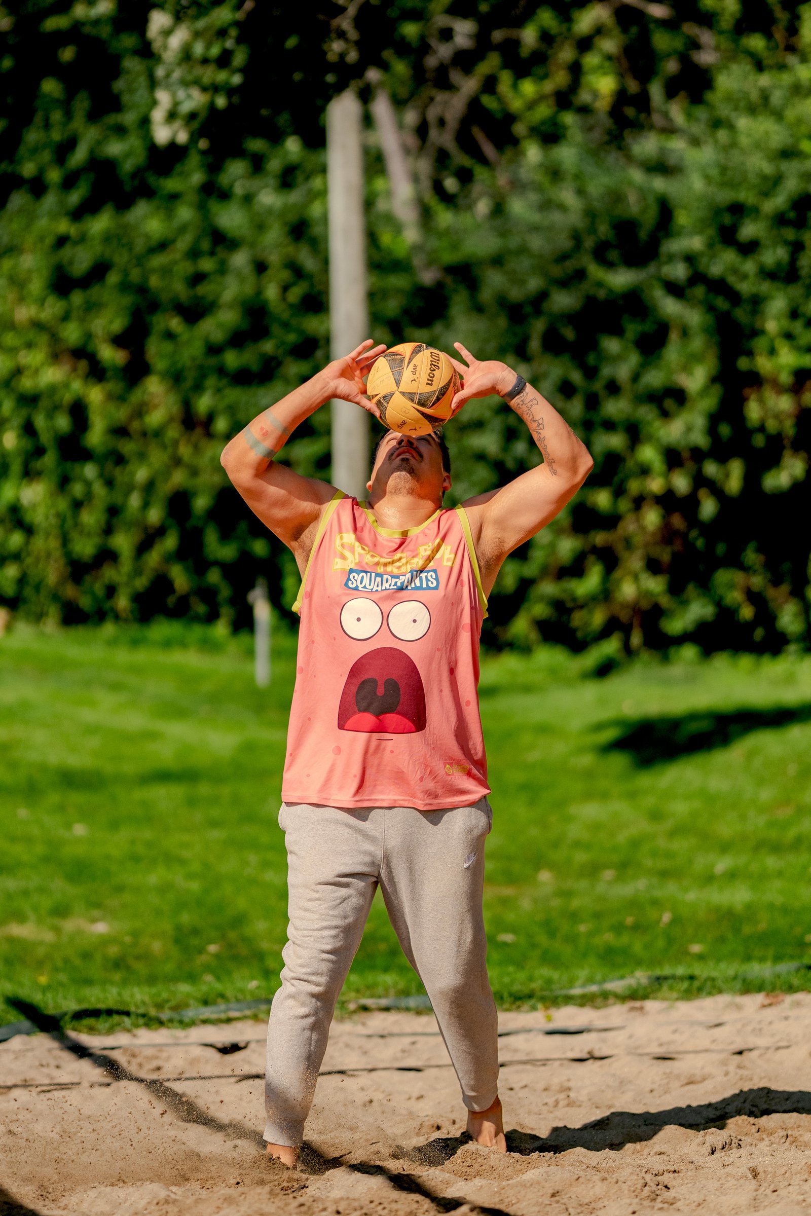 A player in a pink Spongebob Squarepants tank top and grey sweatpants prepares to serve a volleyball on a sand court.