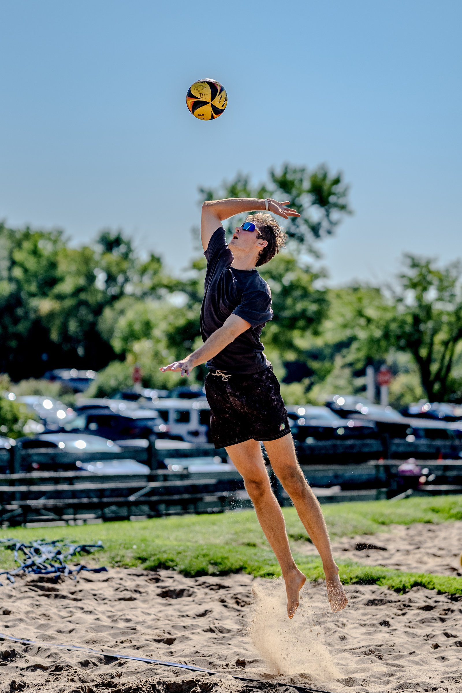 A player in a black shirt and shorts jumps to hit a volleyball on a sandy court with trees and cars in the background.