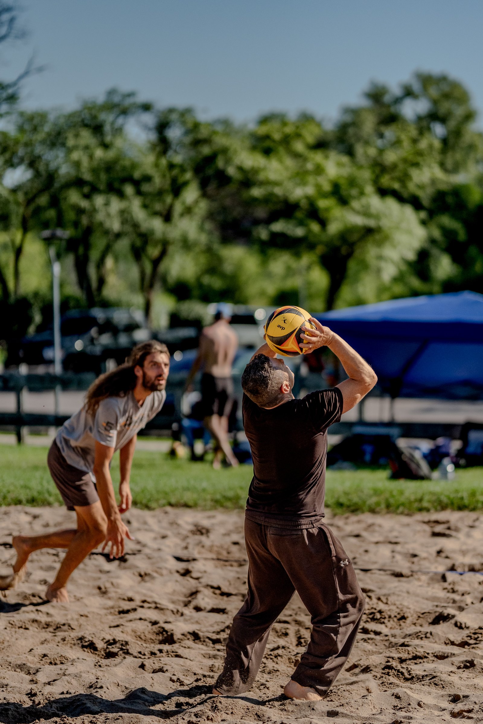 A player in a black top prepares to serve a volleyball on a sand court, with another player in a grey shirt watching.