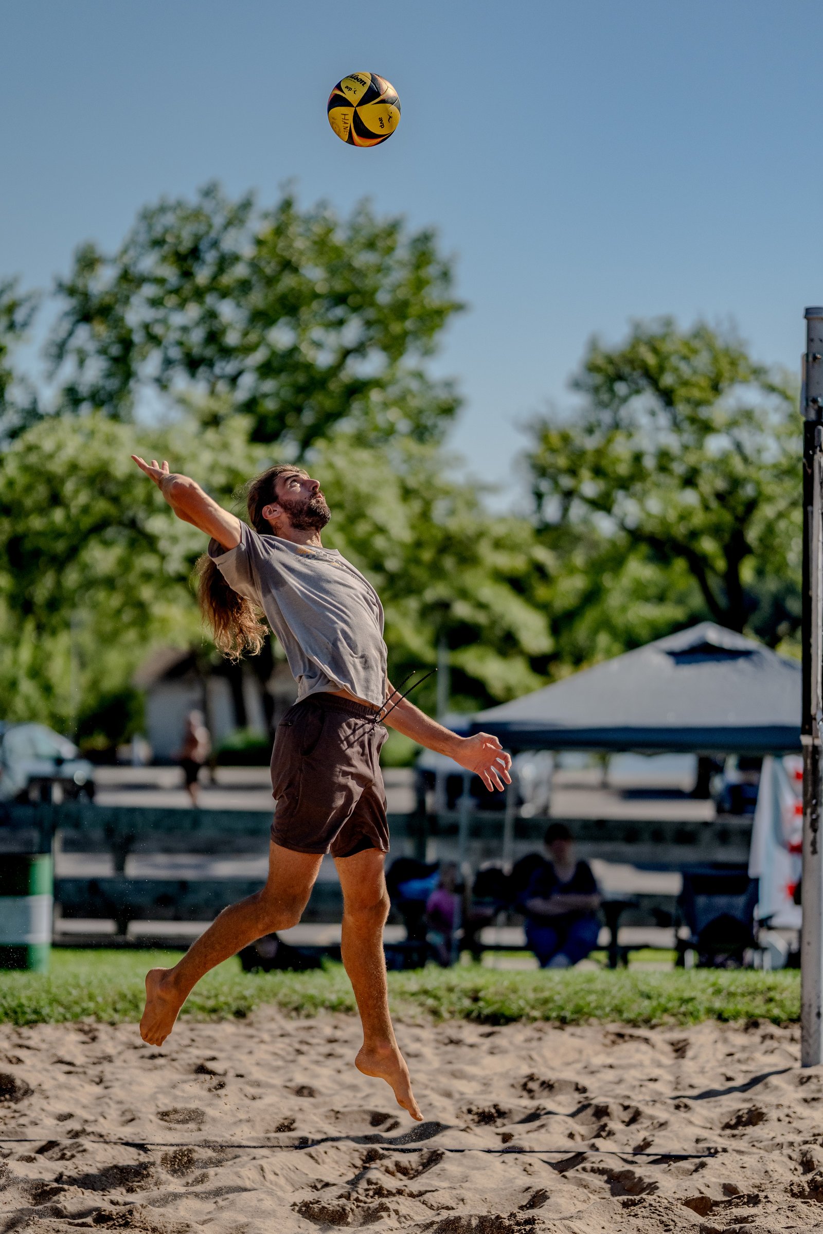 A player in a grey shirt and brown shorts jumps to hit a volleyball on a sandy court with trees and spectators in the background.