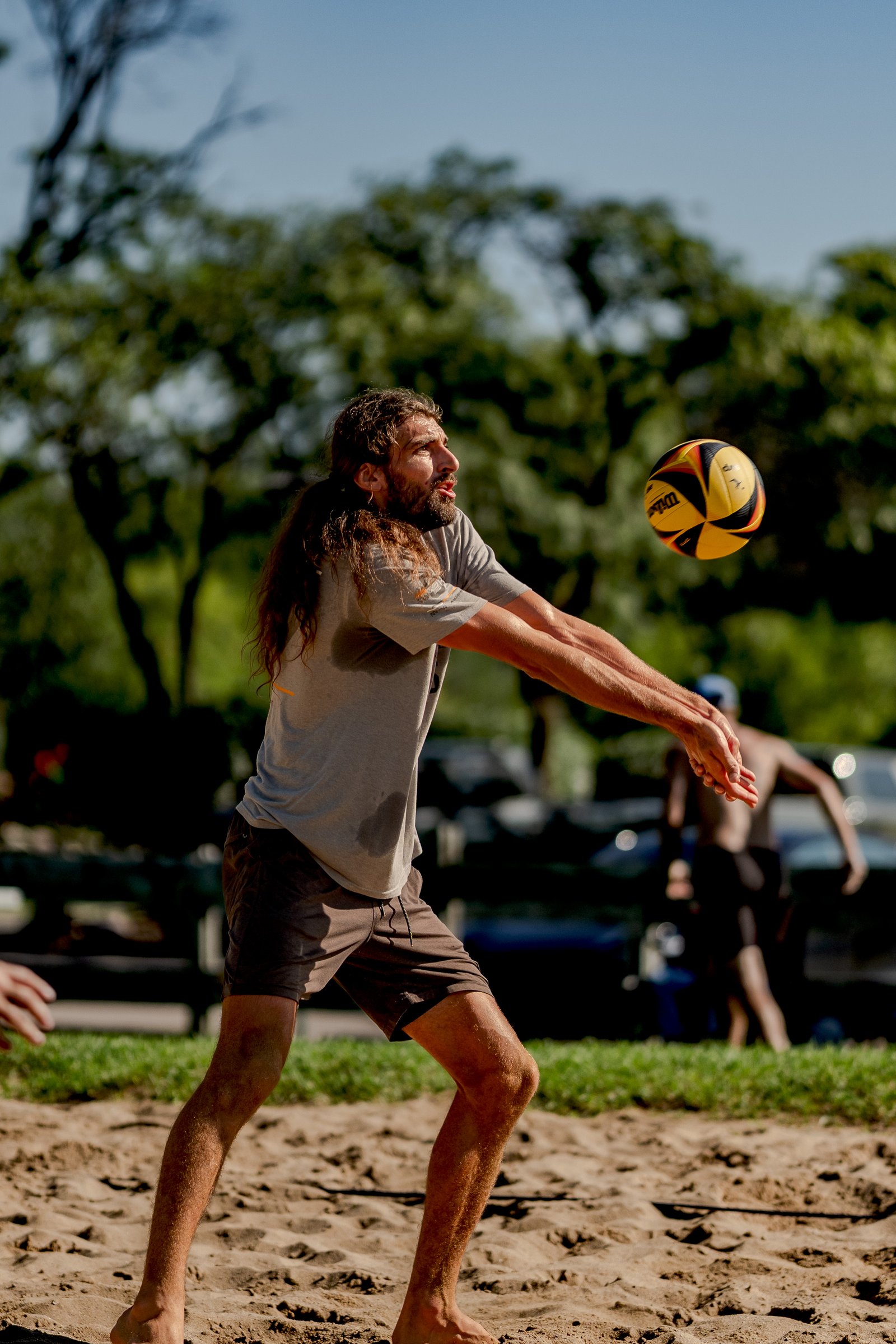 A player in a grey shirt and brown shorts passes a volleyball on a sandy court with trees in the background.
