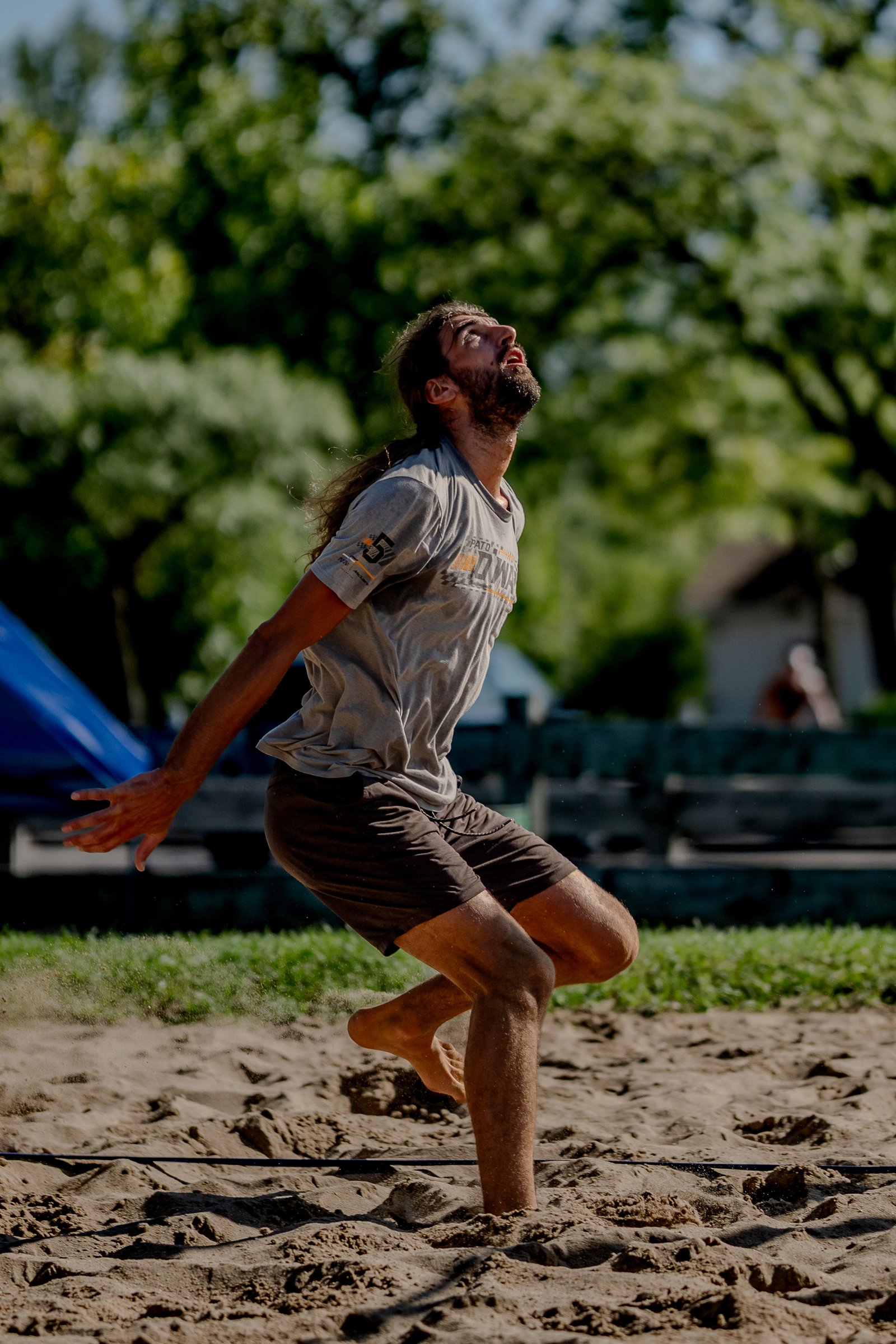 A player in a grey shirt and brown shorts jumps on the sand, looking up.