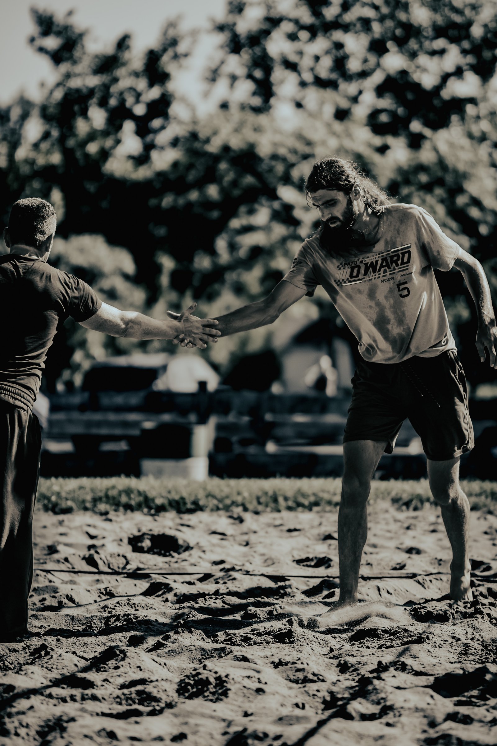 Two players shake hands on a sand court, one in a light-colored shirt with number 5 and the other in a dark shirt.