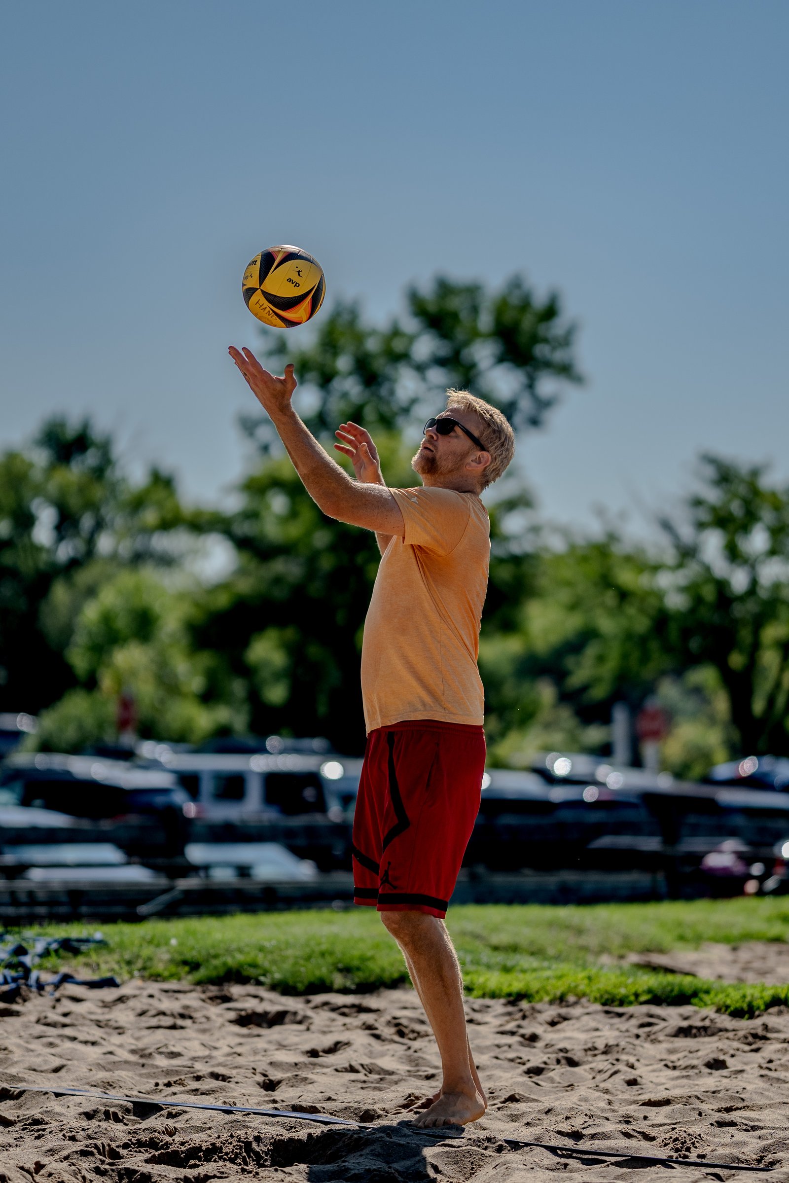A player in an orange top and red shorts tosses a volleyball on a sand court with trees and cars in the background.