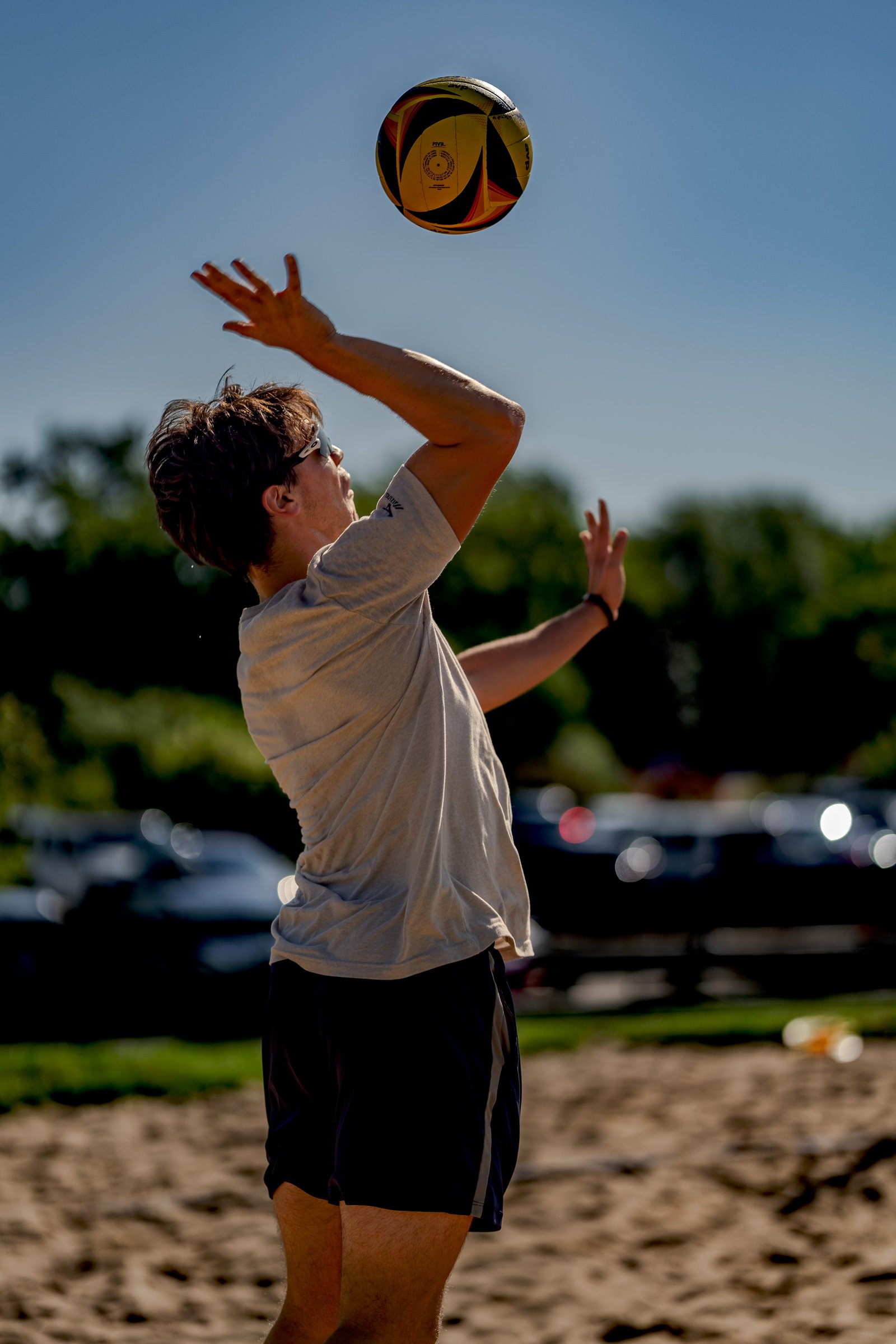 A player in a grey shirt and black shorts serves a volleyball on a sandy court.