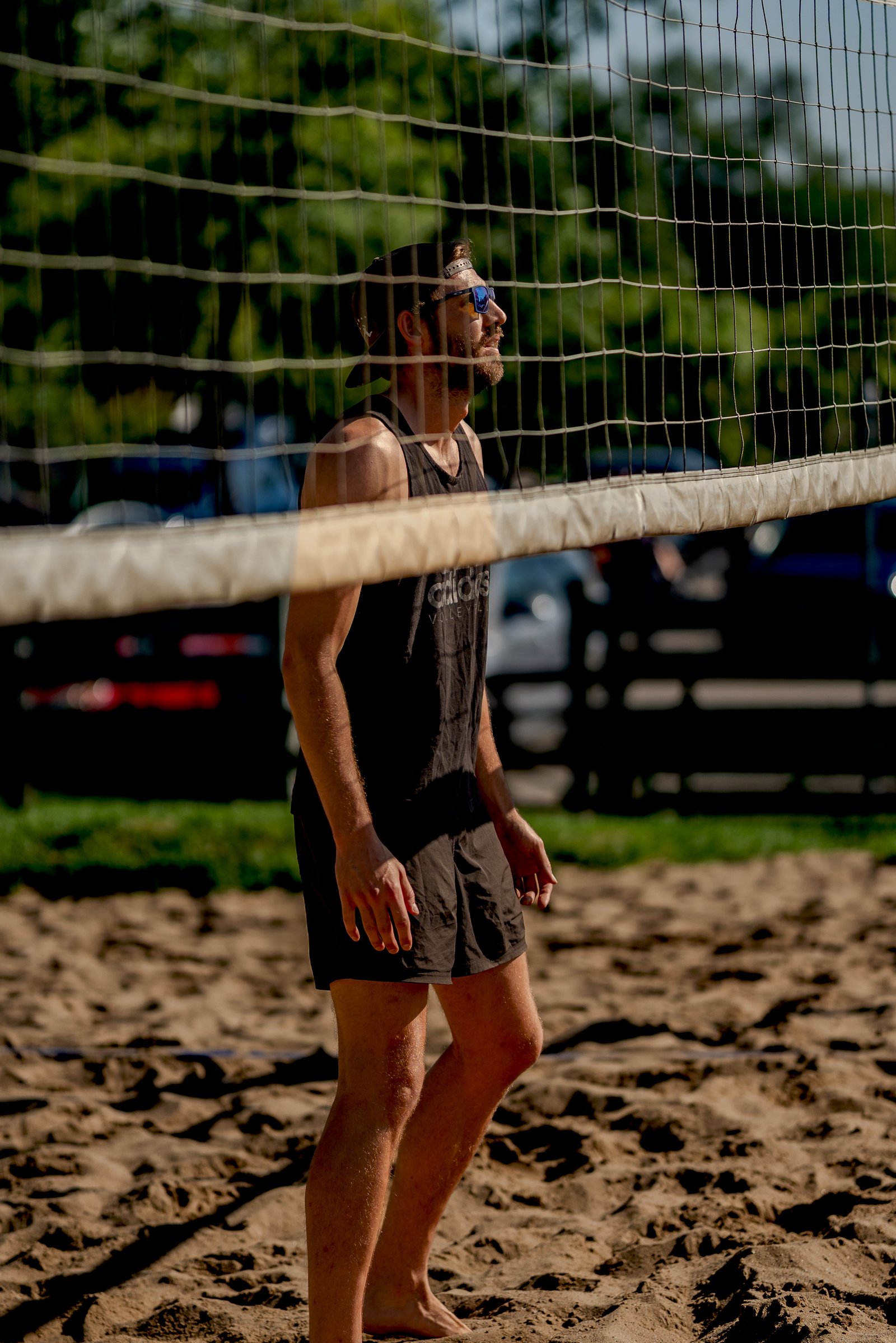 A player in a black tank top and shorts stands on the sand near a volleyball net, looking to the side.