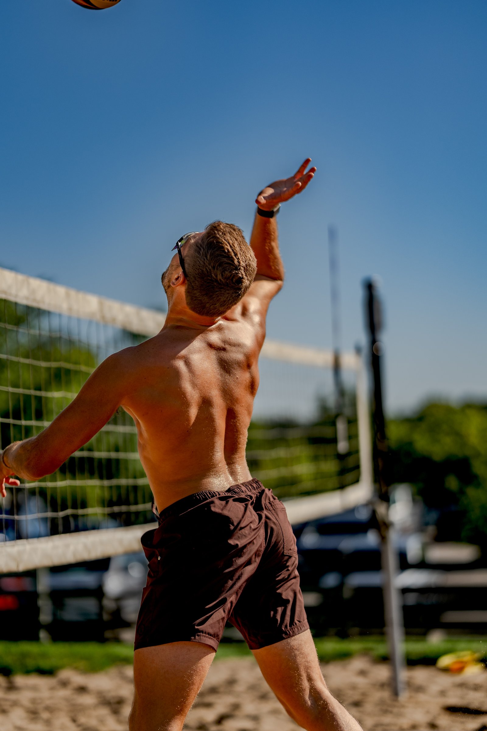 A player in brown shorts serves a volleyball on a sandy court with a net and trees in the background.