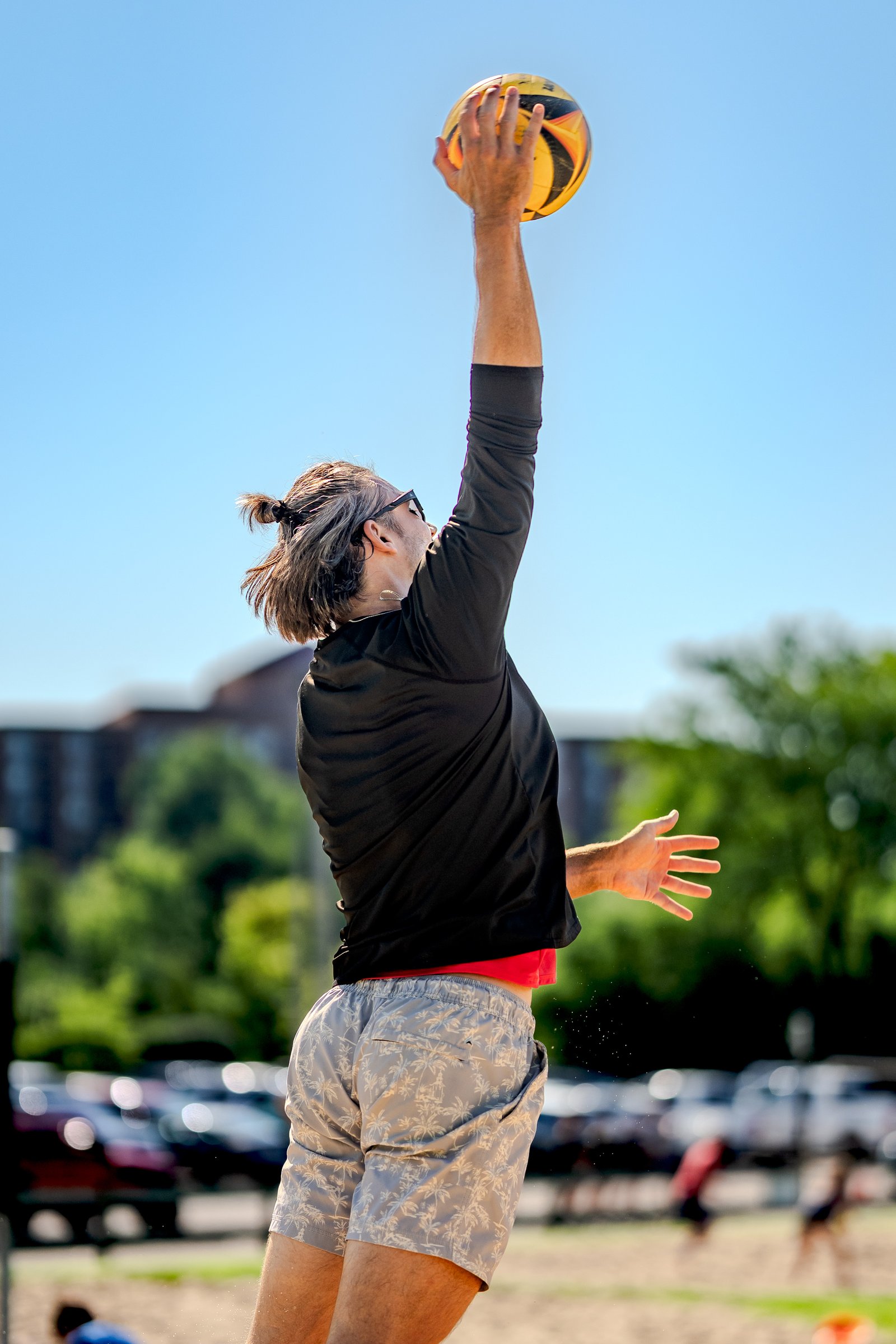 A player in a black top and patterned shorts reaches up to hit a volleyball during a tournament.