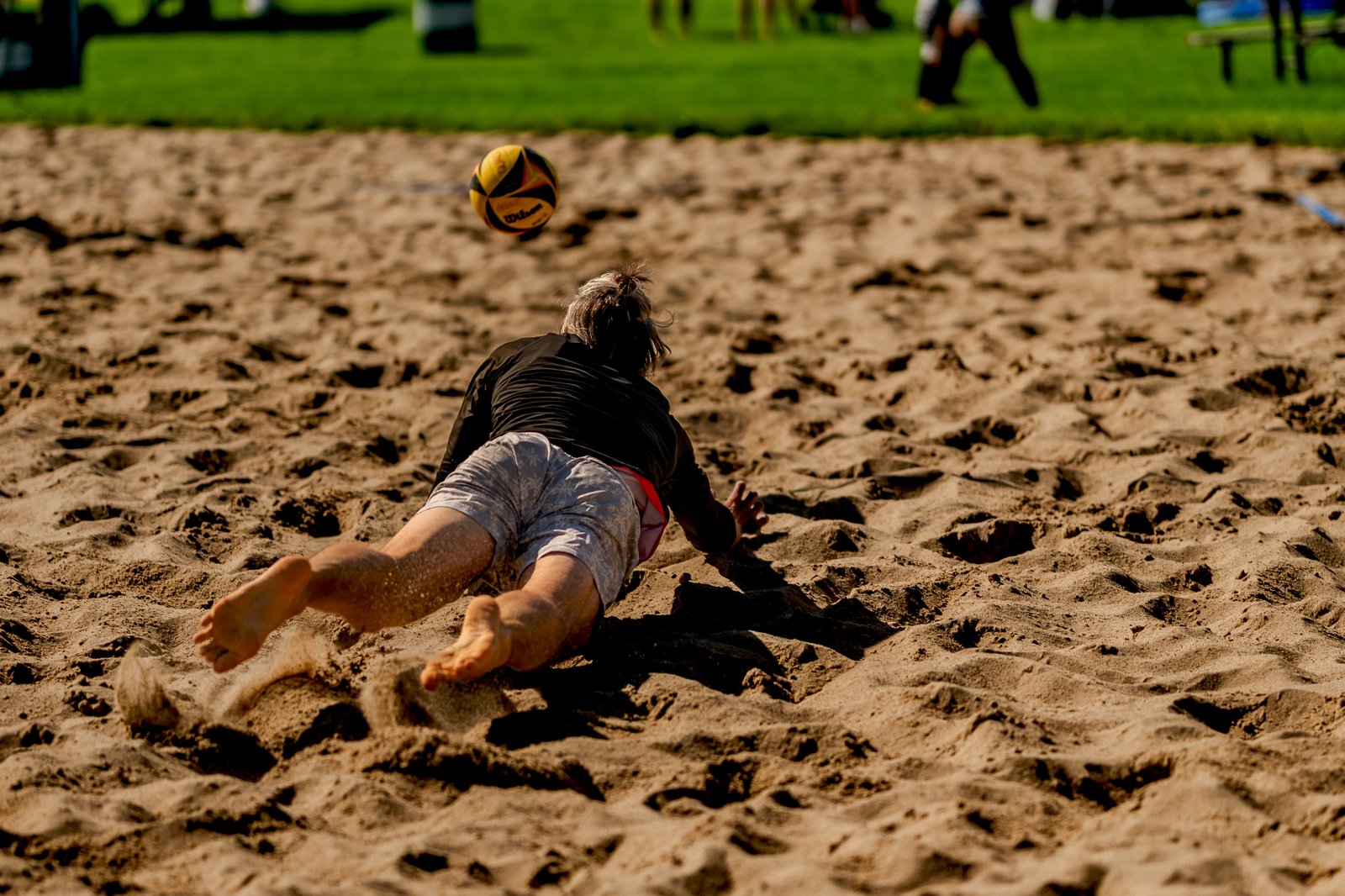 A player in a black top and grey shorts dives for a volleyball on a sand court.