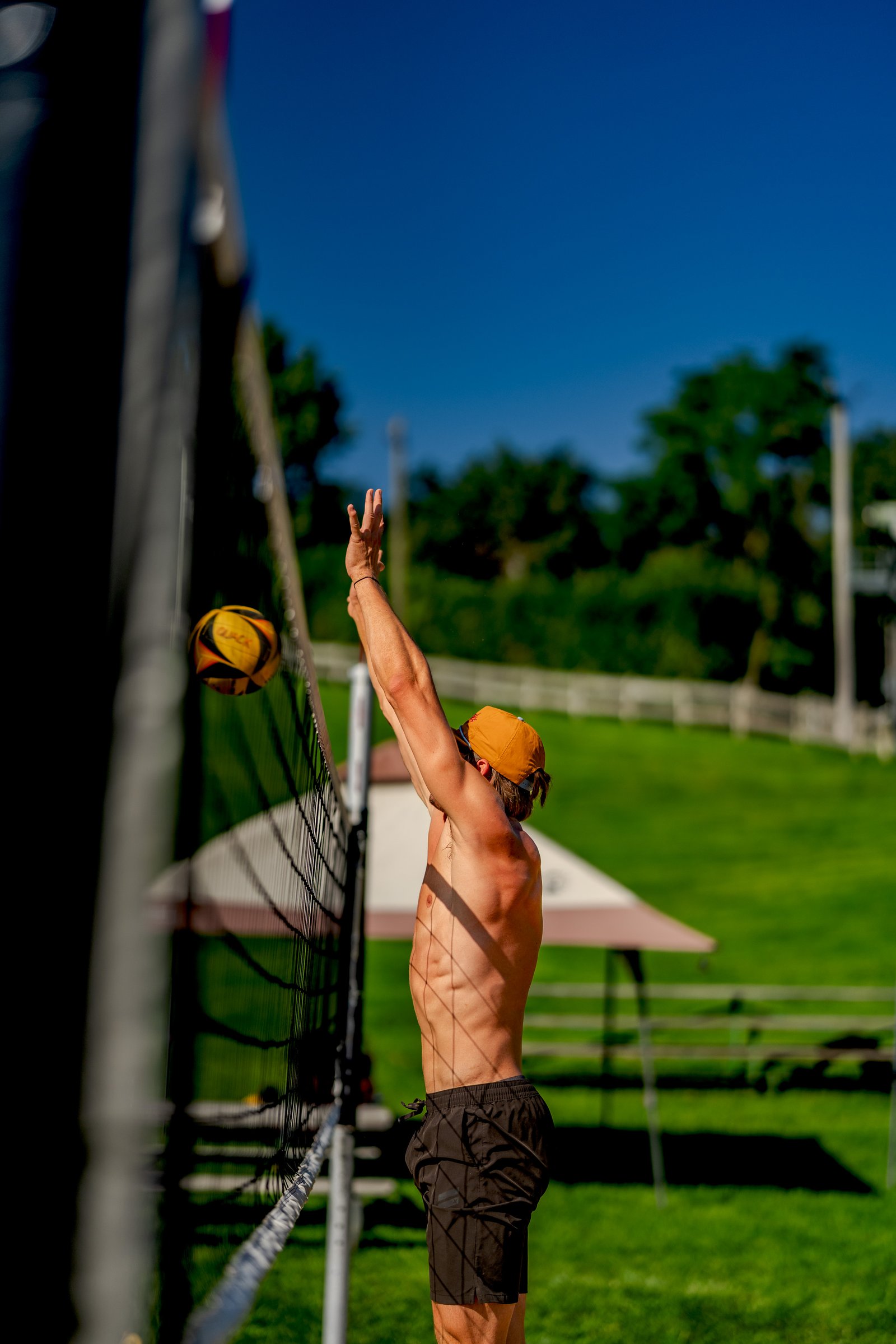 A player in black shorts and an orange cap prepares to serve a volleyball over the net on a sunny day.