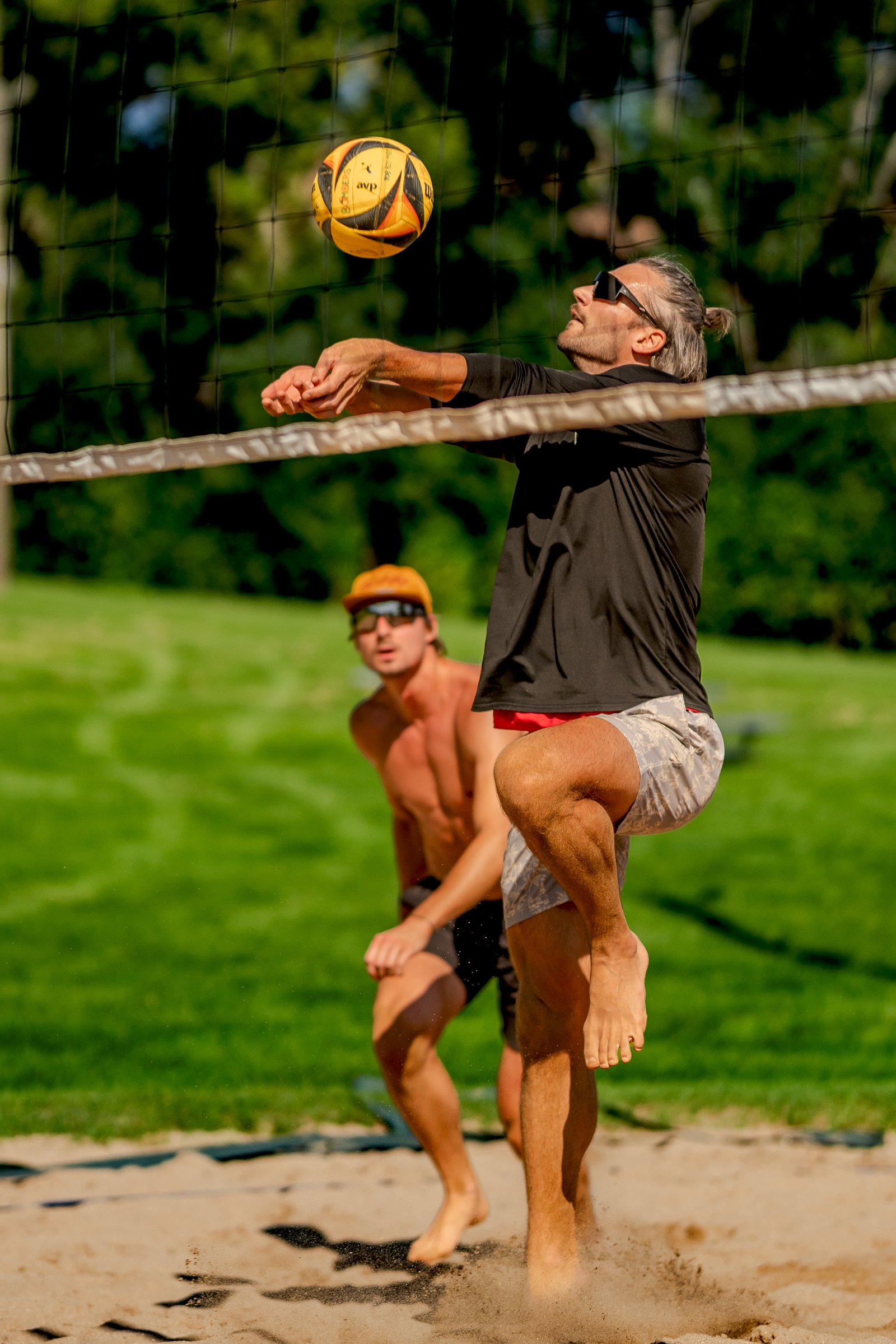 A player in a black top and patterned shorts jumps to hit a volleyball, with another player in orange shorts and a hat watching.