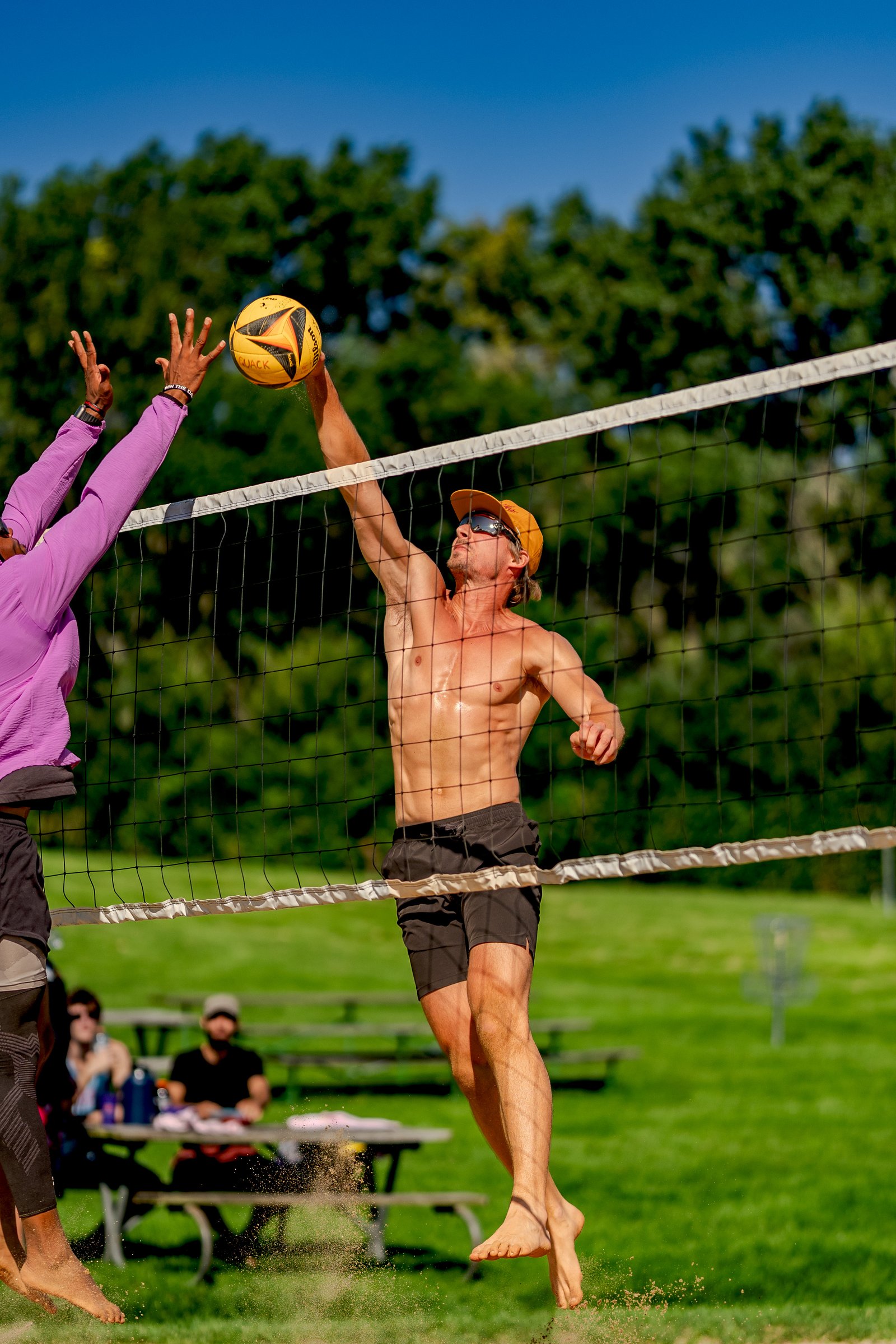 A player in a purple top reaches for the ball while a player in black shorts jumps to hit it over the net.