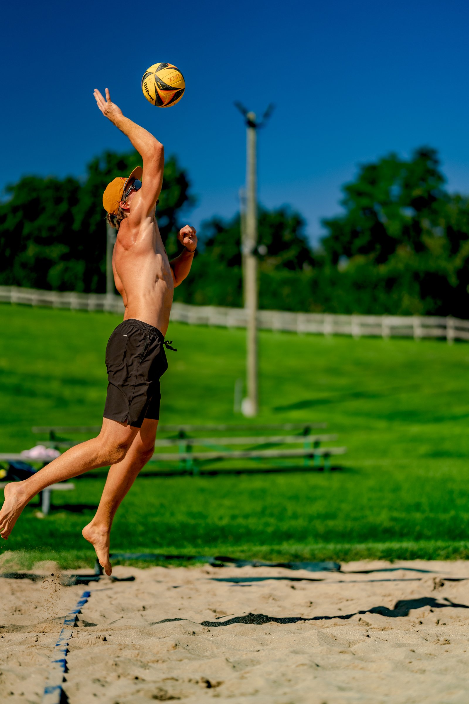 A player in black shorts and an orange hat serves a volleyball on a sand court with a grassy background.