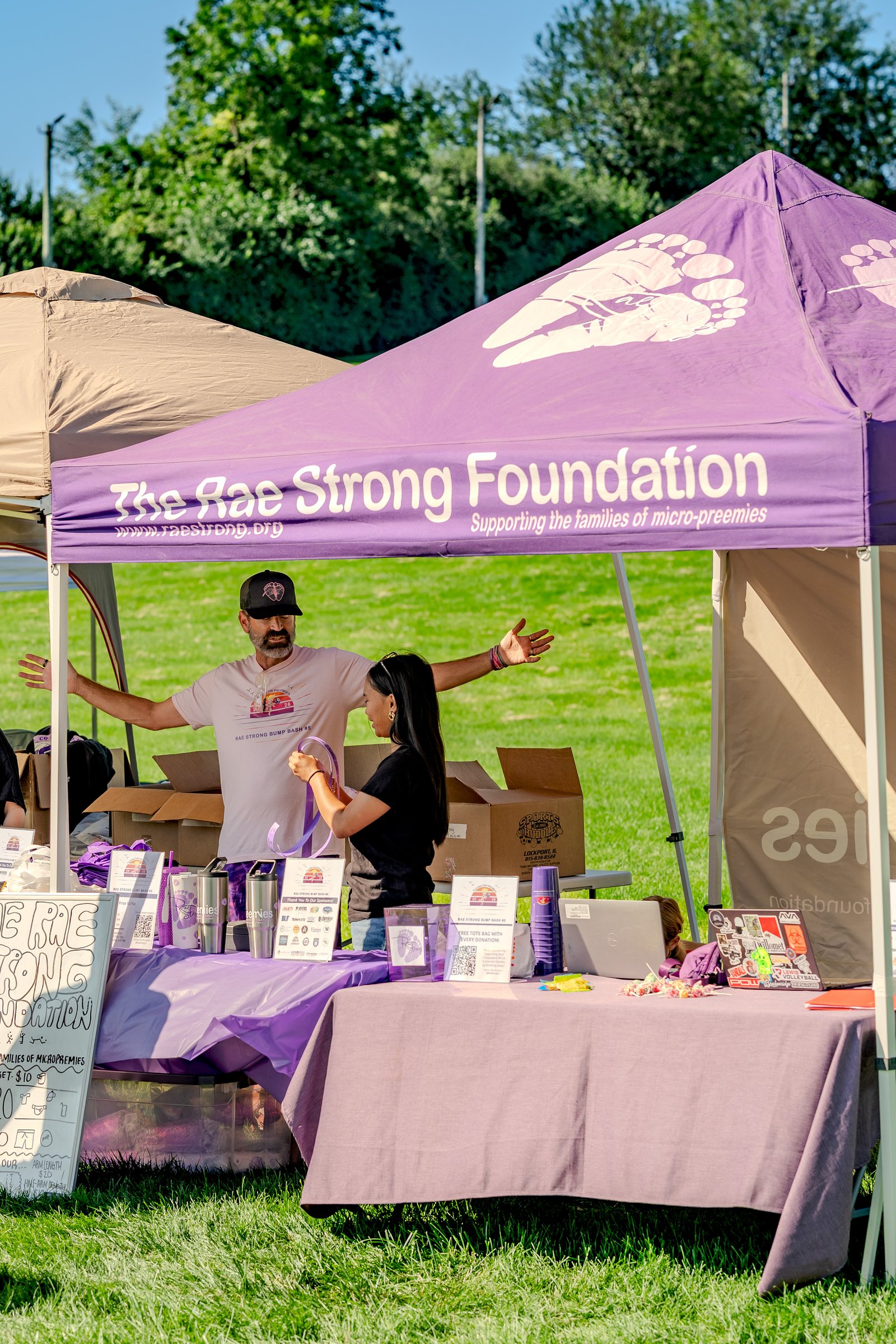 A man in a pink shirt and a woman in a black top stand behind a table at a charity event with a purple tent.