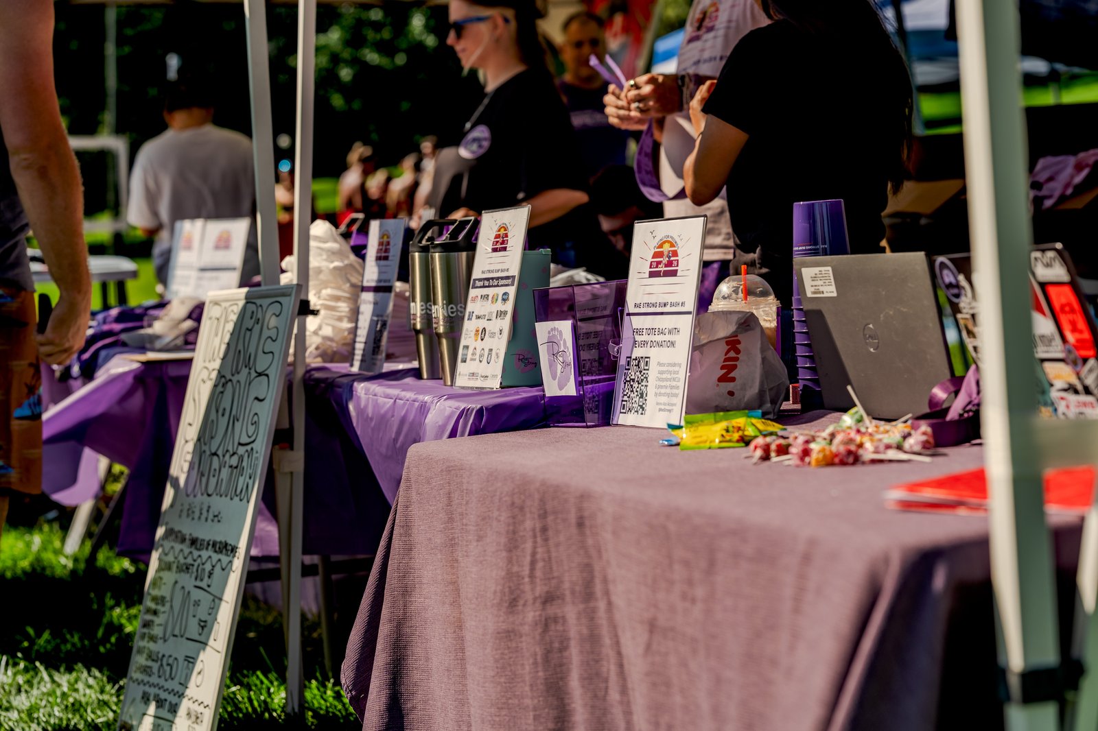 A player in a black top and a player in purple shorts are visible at a table with various items, including a laptop and candy, during a charity volleyball tournament.