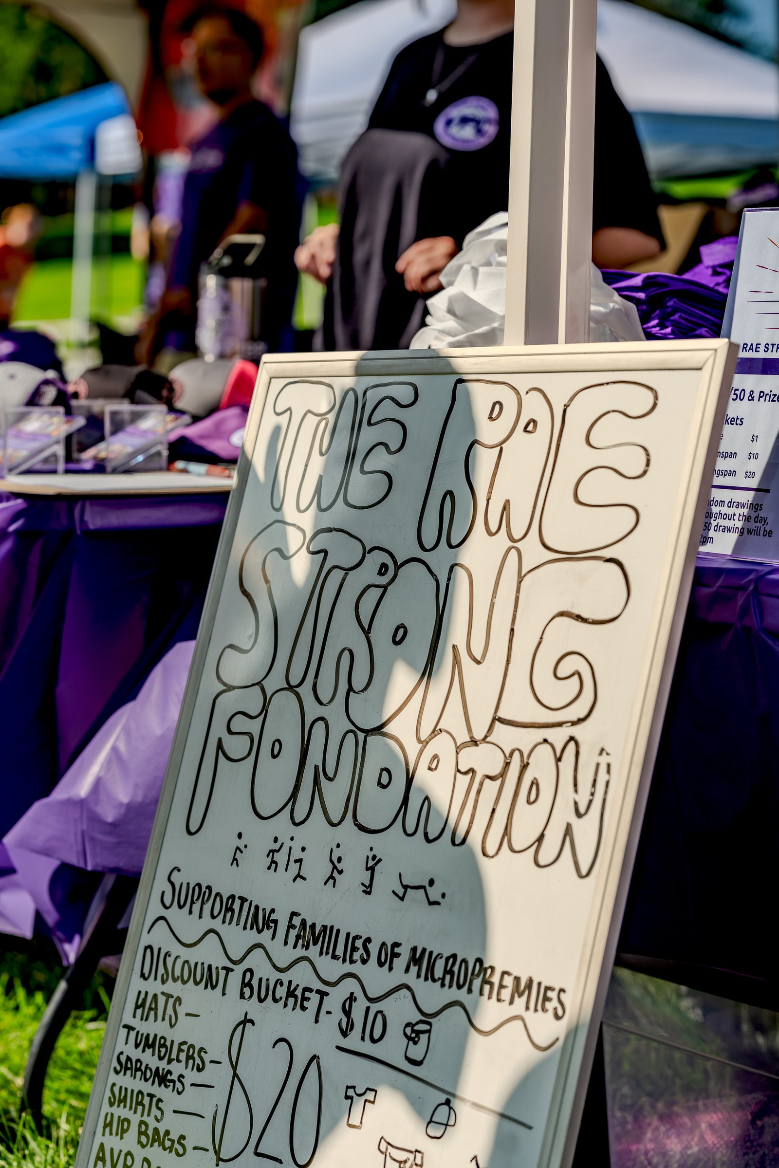 A sign for The Rae Strong Foundation displays prices for hats, tumblers, sarongs, shirts, and hip bags, with a discount bucket for $10 and items for $20.