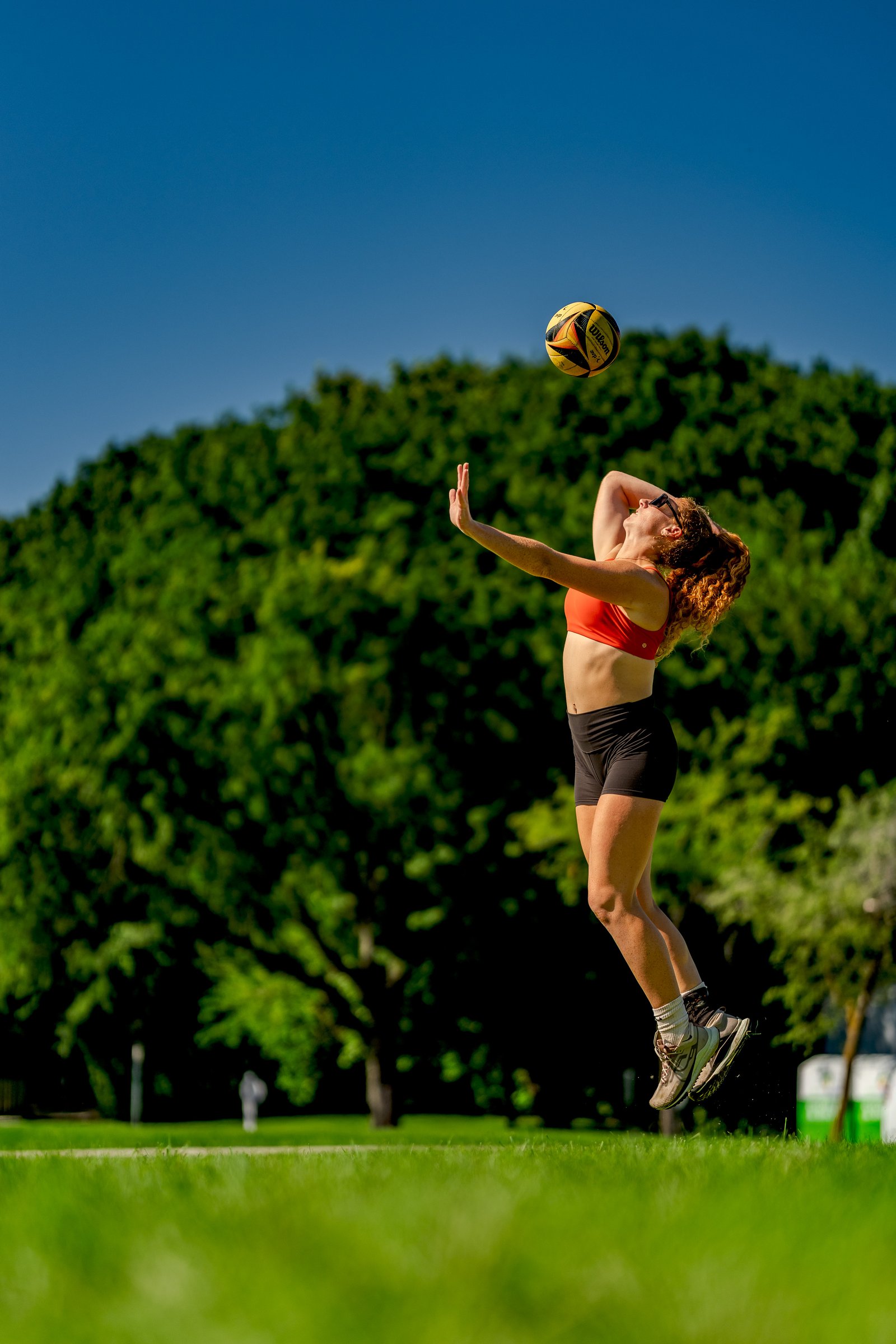 A player in an orange top and black shorts jumps to hit a volleyball against a green tree backdrop.