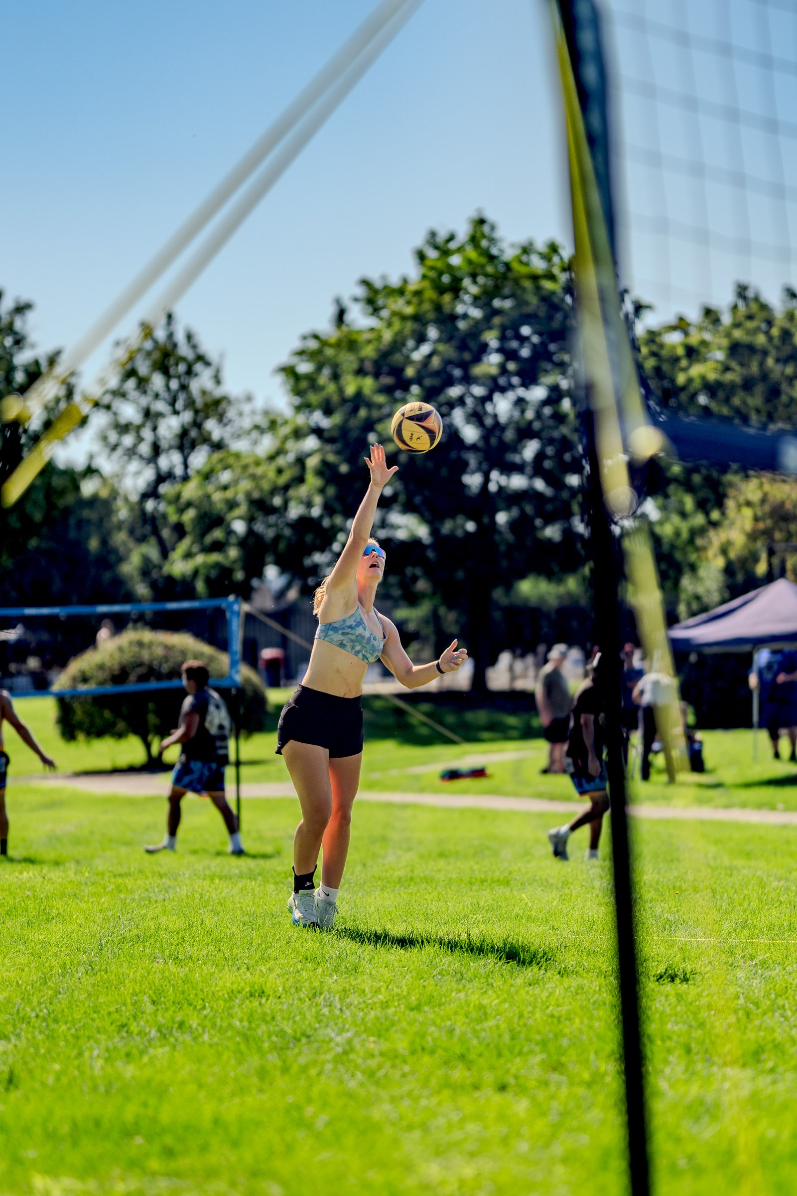 A player in a camouflage top and black shorts reaches for the ball during a volleyball game on a sunny day.