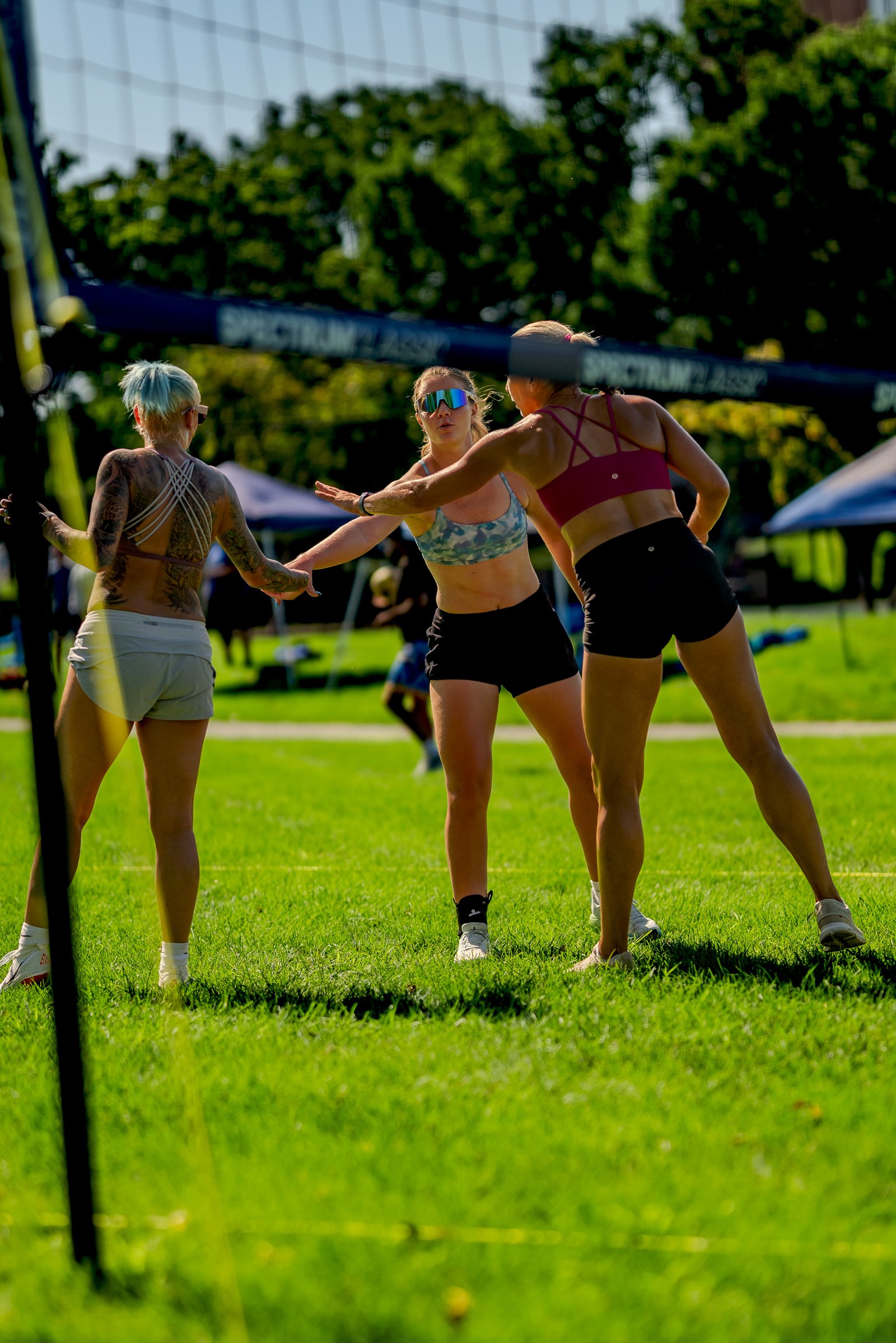 Players in black shorts and patterned tops reach for the ball during a volleyball match on a grassy field.
