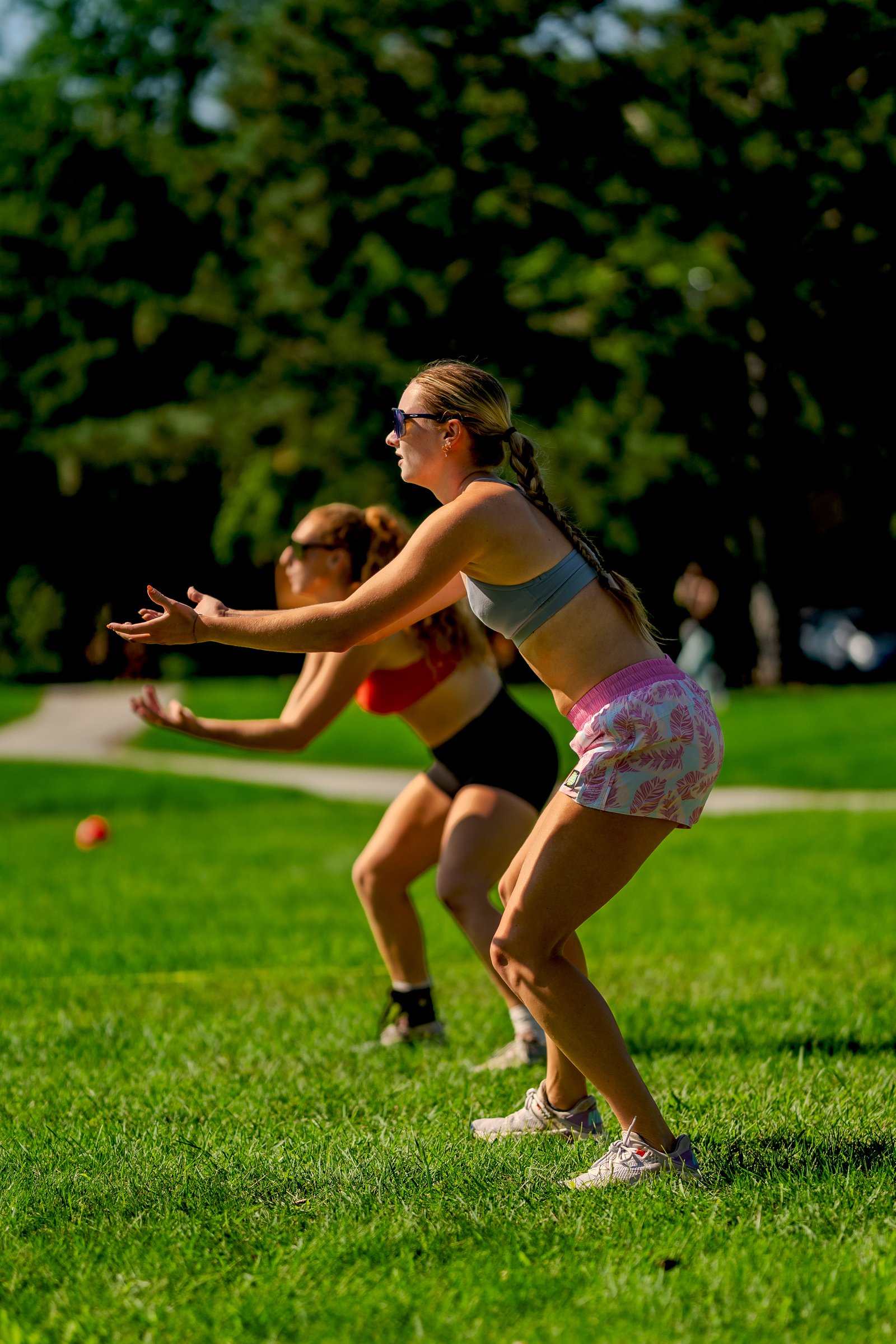 A player in a gray top and pink shorts prepares to receive a volleyball, with another player in a red top behind her on the grass.