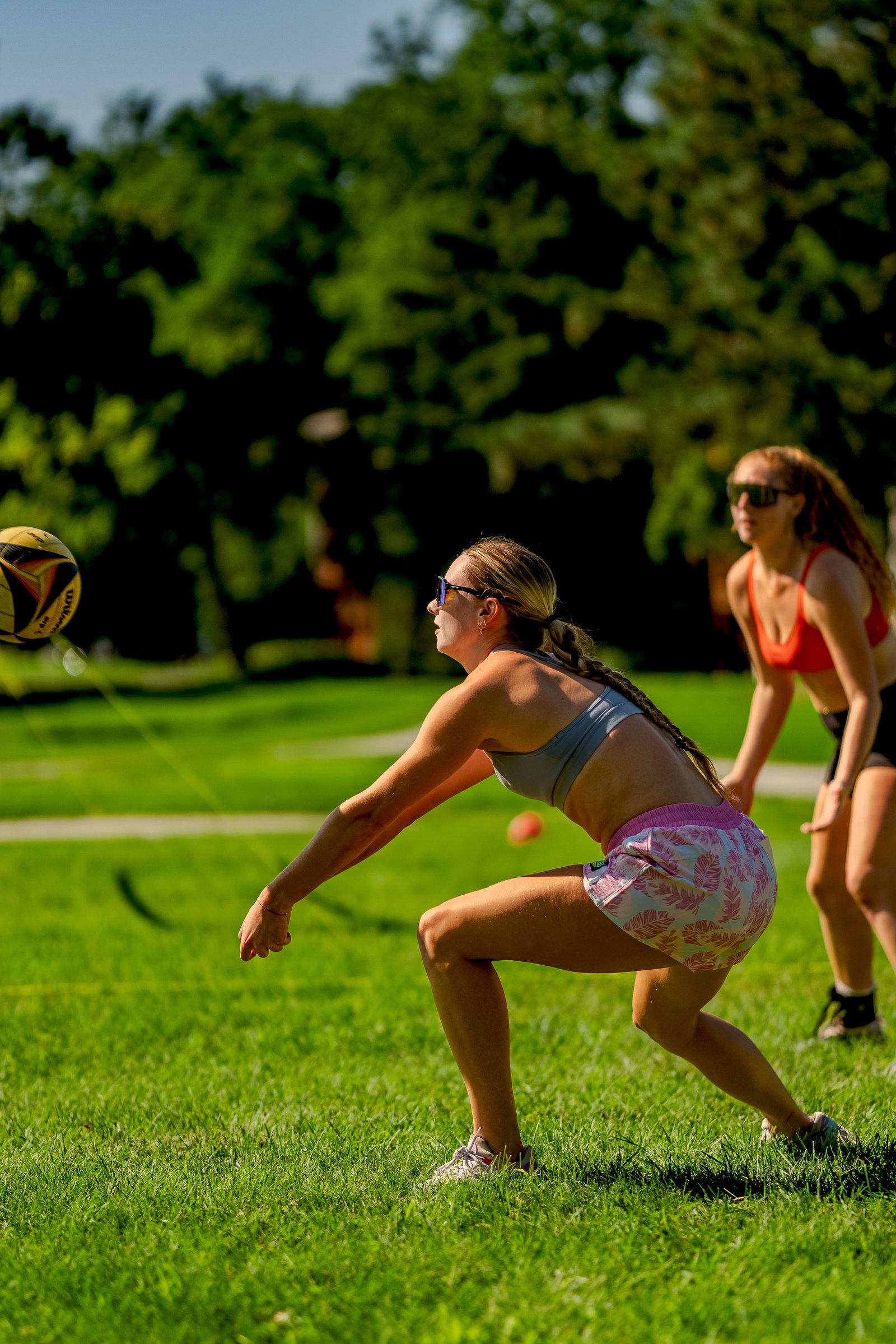 A player in a grey top and patterned shorts prepares to pass a volleyball, with another player in a red top behind her on the grass.