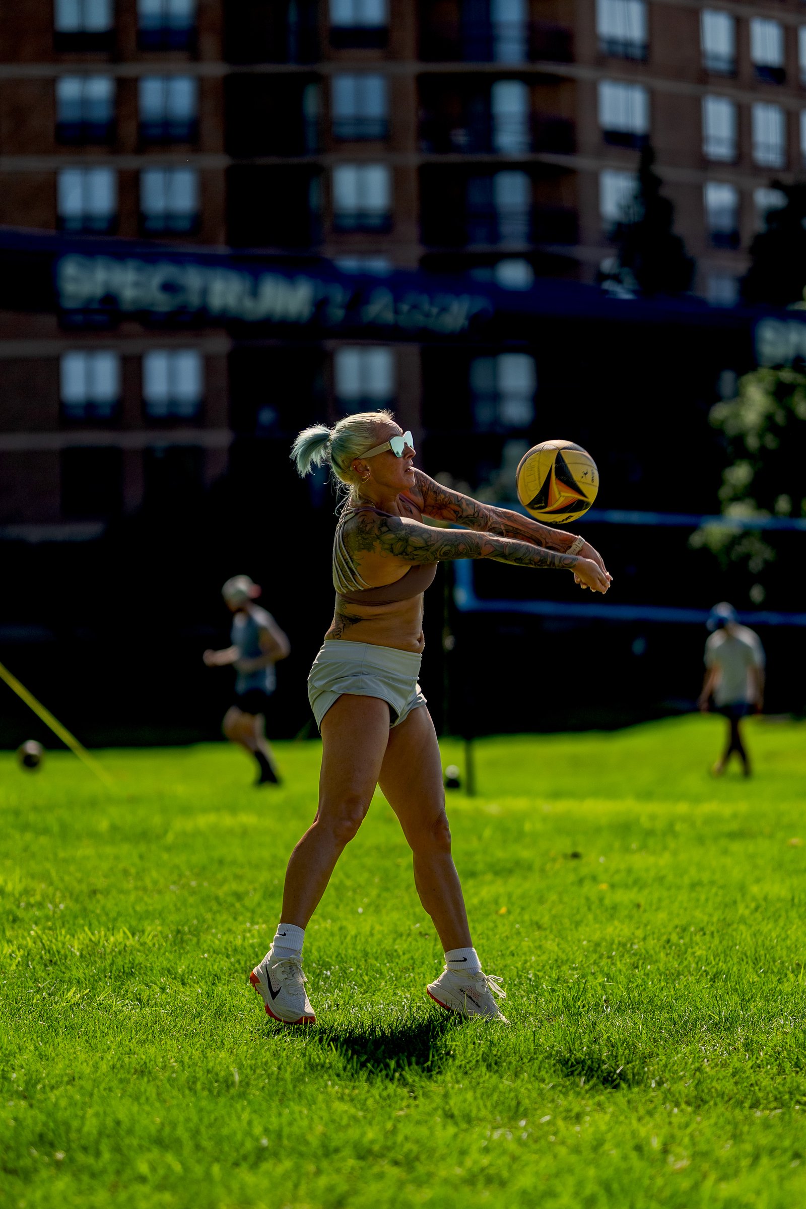 A player in a brown top and grey shorts passes a volleyball on a grassy court with a building in the background.