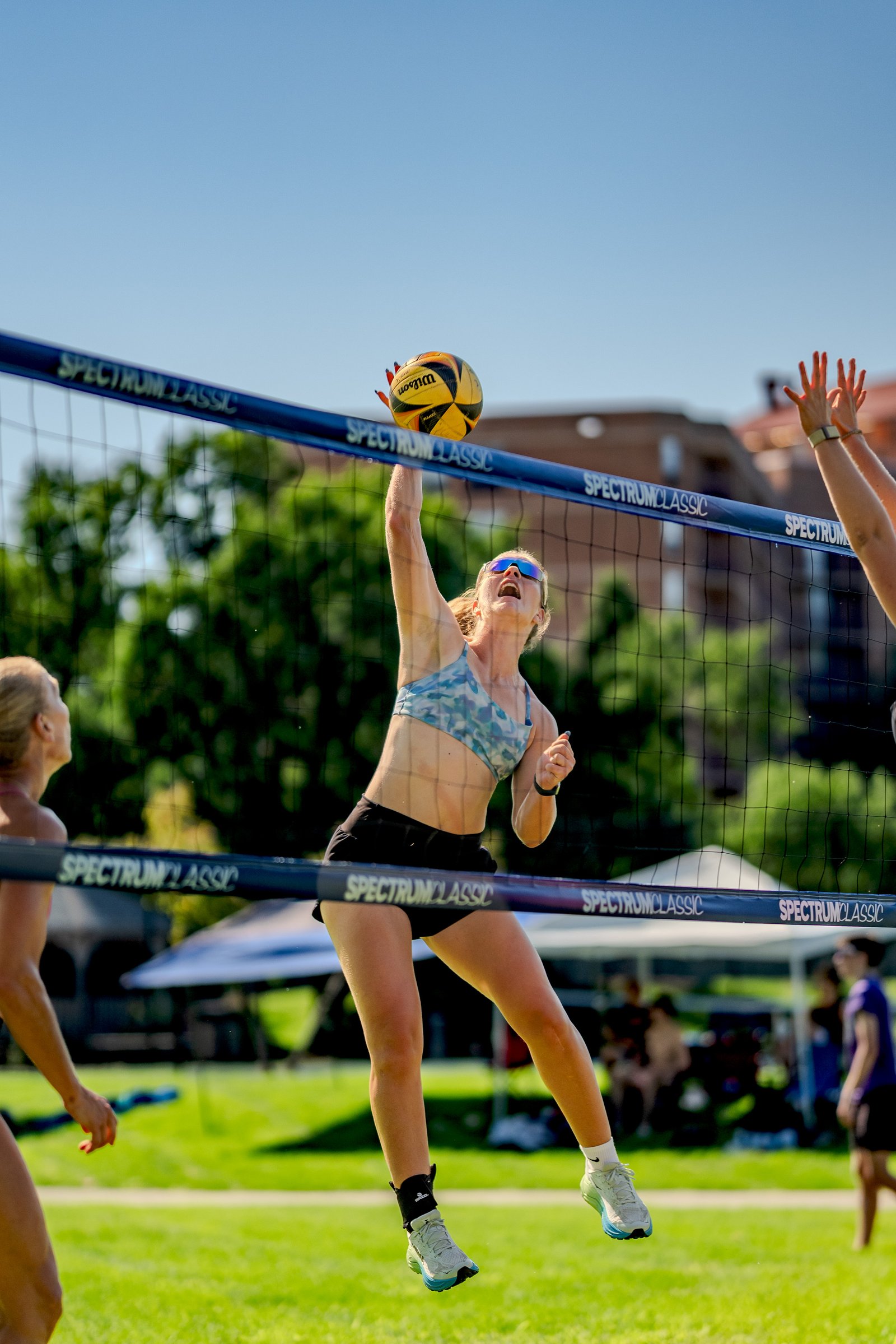 A player in a blue and white patterned top jumps to hit the volleyball over the net, with another player in the background reaching up.