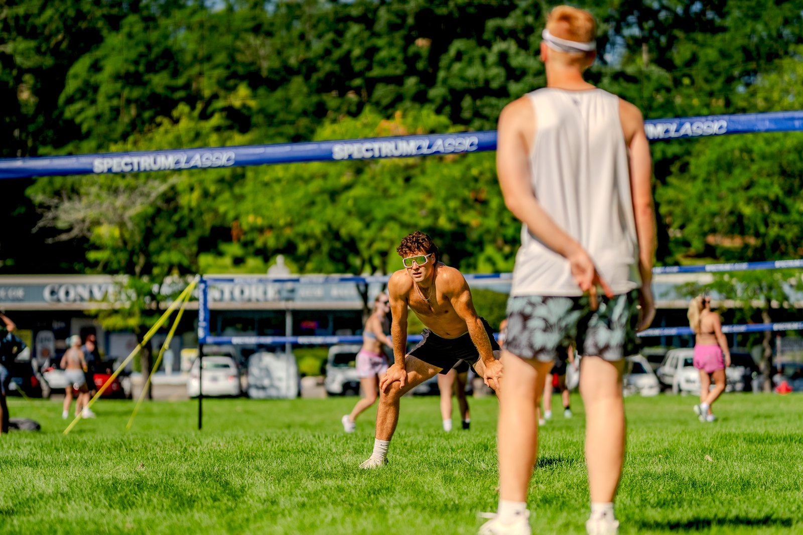 A player in black shorts crouches on the grass, while another player in a white top stands with hands behind their back during a volleyball match.