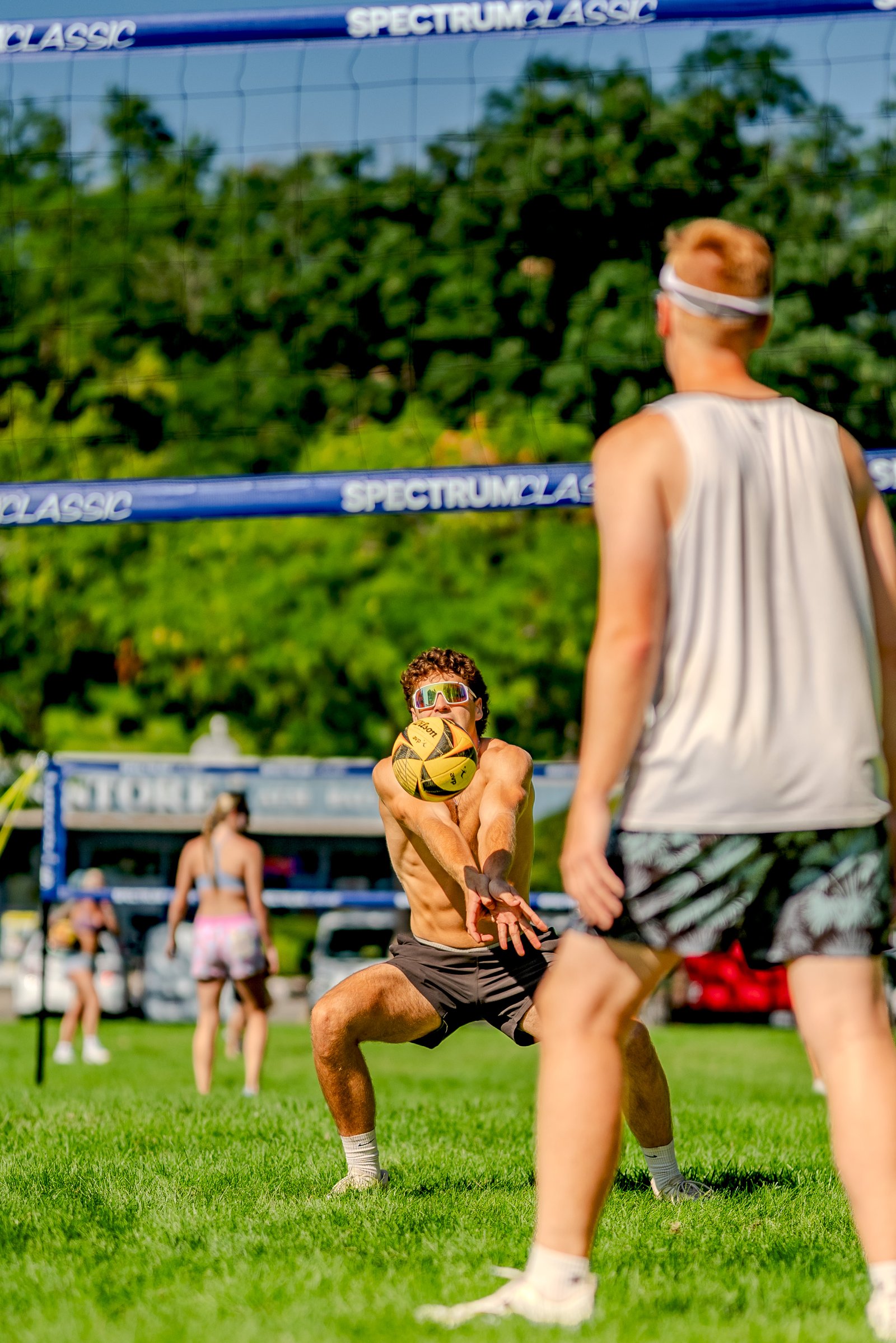 A player in black shorts passes a volleyball while another player in a white top watches on a sunny grass court.