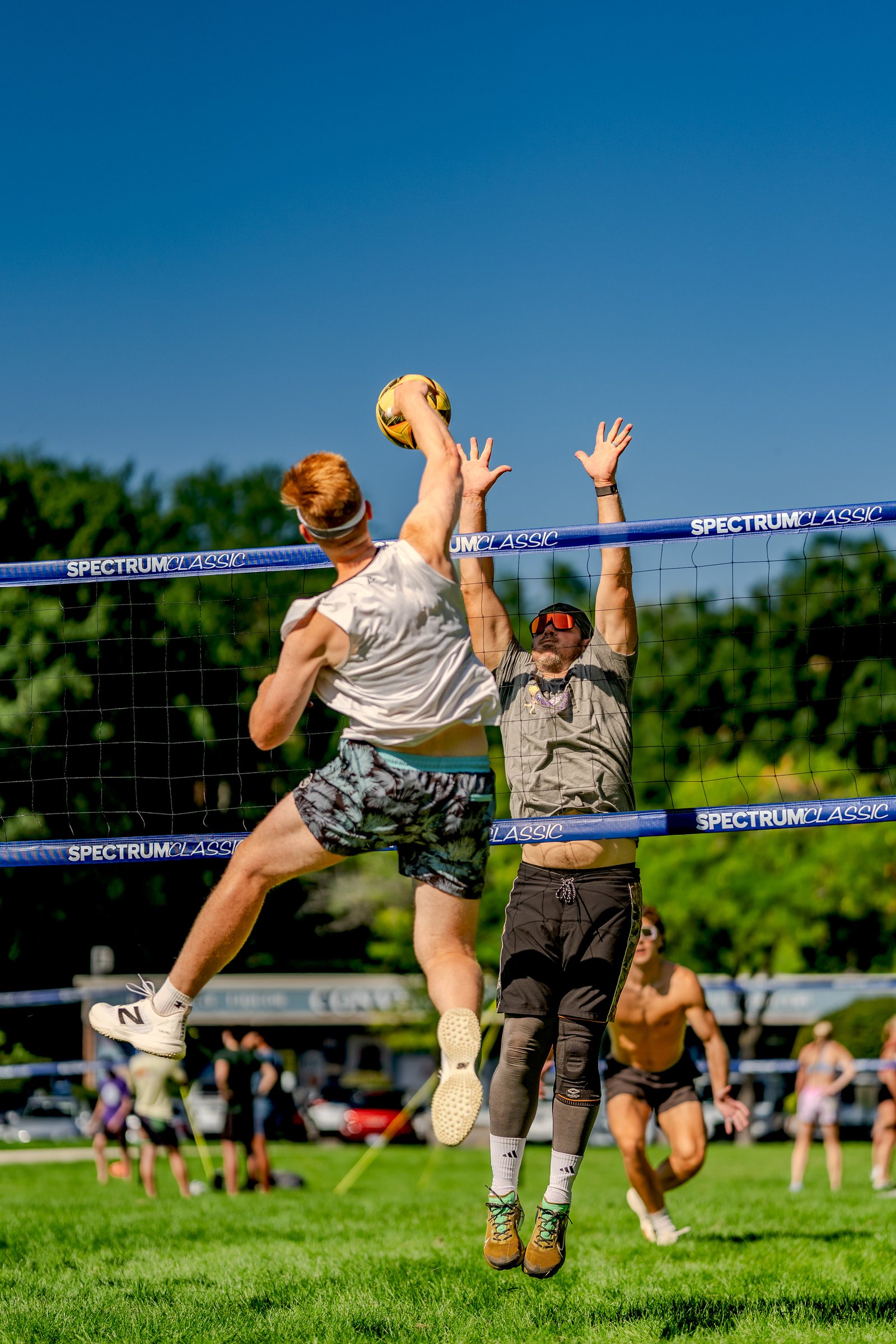 A player in a white top jumps to hit the ball over the net while another player in a gray top reaches up to block during a volleyball game.